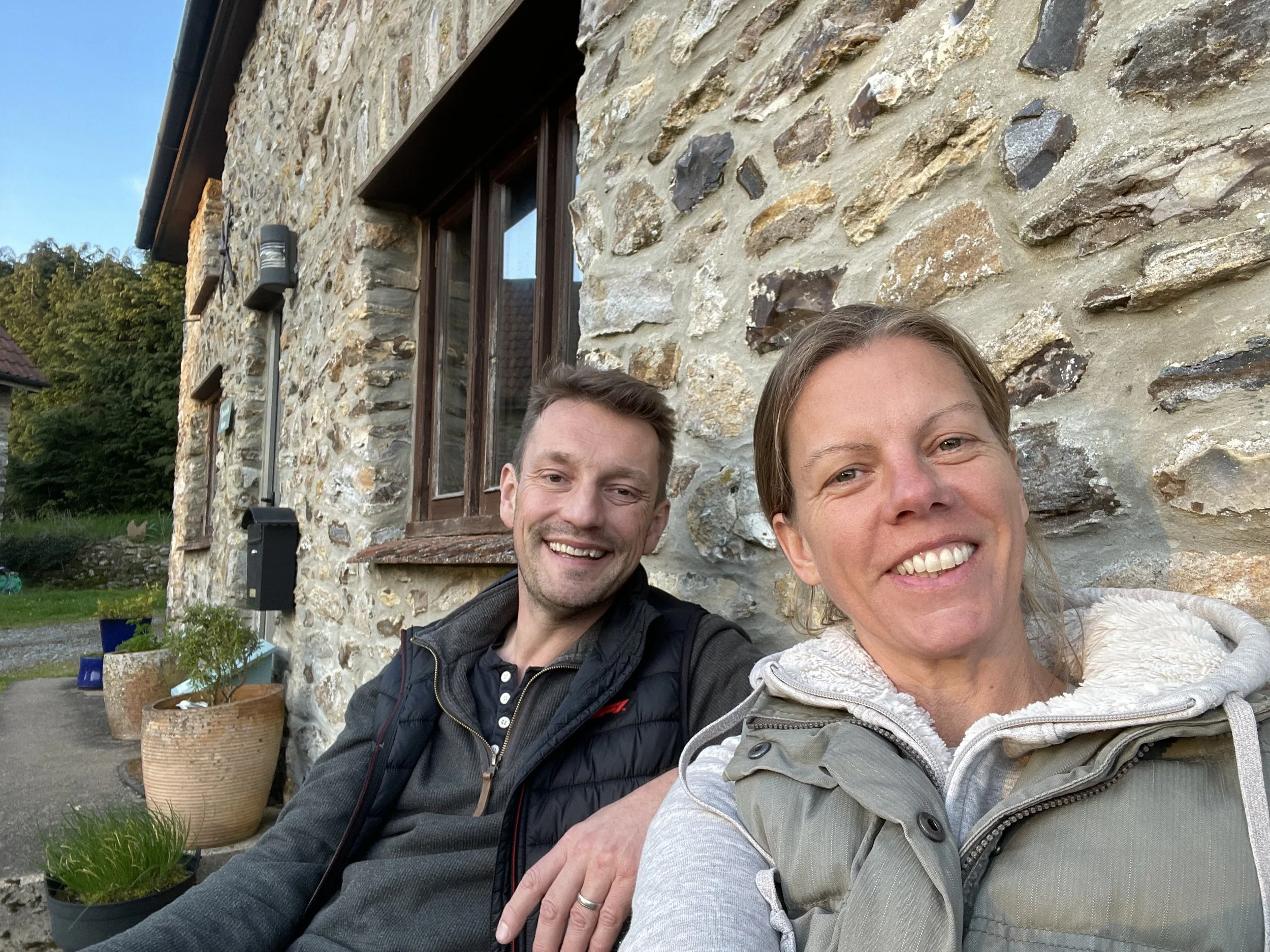 Two smiling people, James and Natasha, taking a selfie in front of a stone building with a window and some plants in pots nearby.