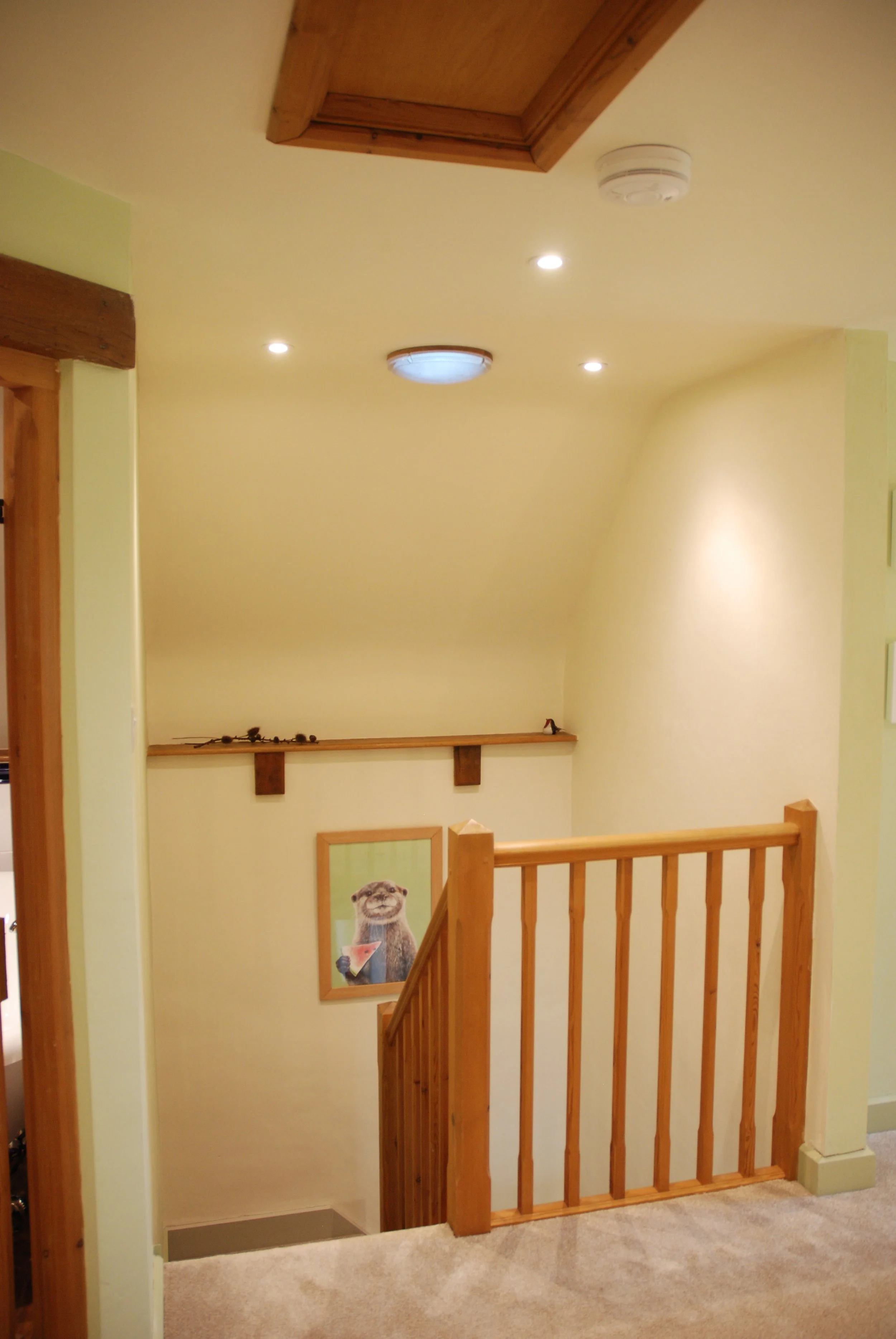 Interior view of a staircase area with wooden railing, a picture of a beaver holding a drink on the wall, a plastic owl on the ledge, and a ceiling hatch with lights.
