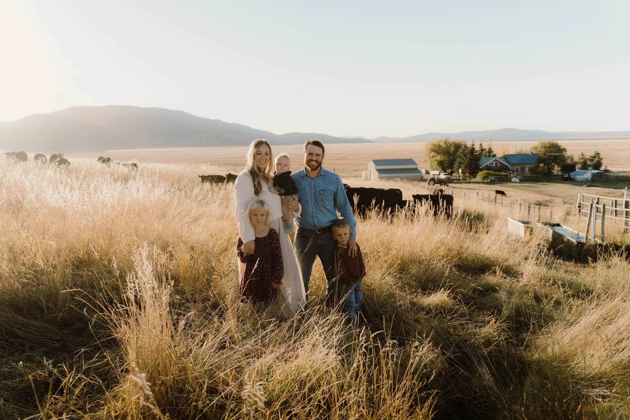 Family of five, including two adults and three children, standing in a grassy field with cattle and farm buildings in the background at sunset.