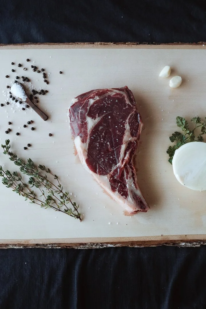 Raw beef steak on a wooden cutting board, seasoned with salt and pepper, with garlic cloves, fresh herbs, and a dollop of cheese around it.