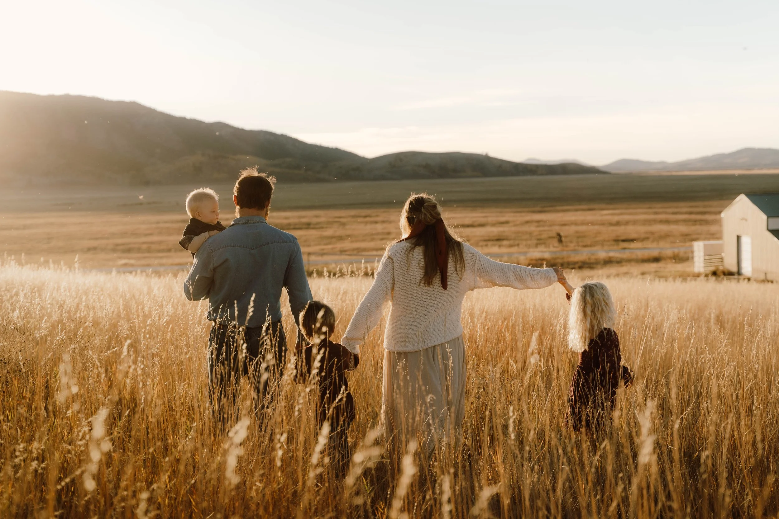 A family of five walking through a golden wheat field at sunset, with mountains in the background.