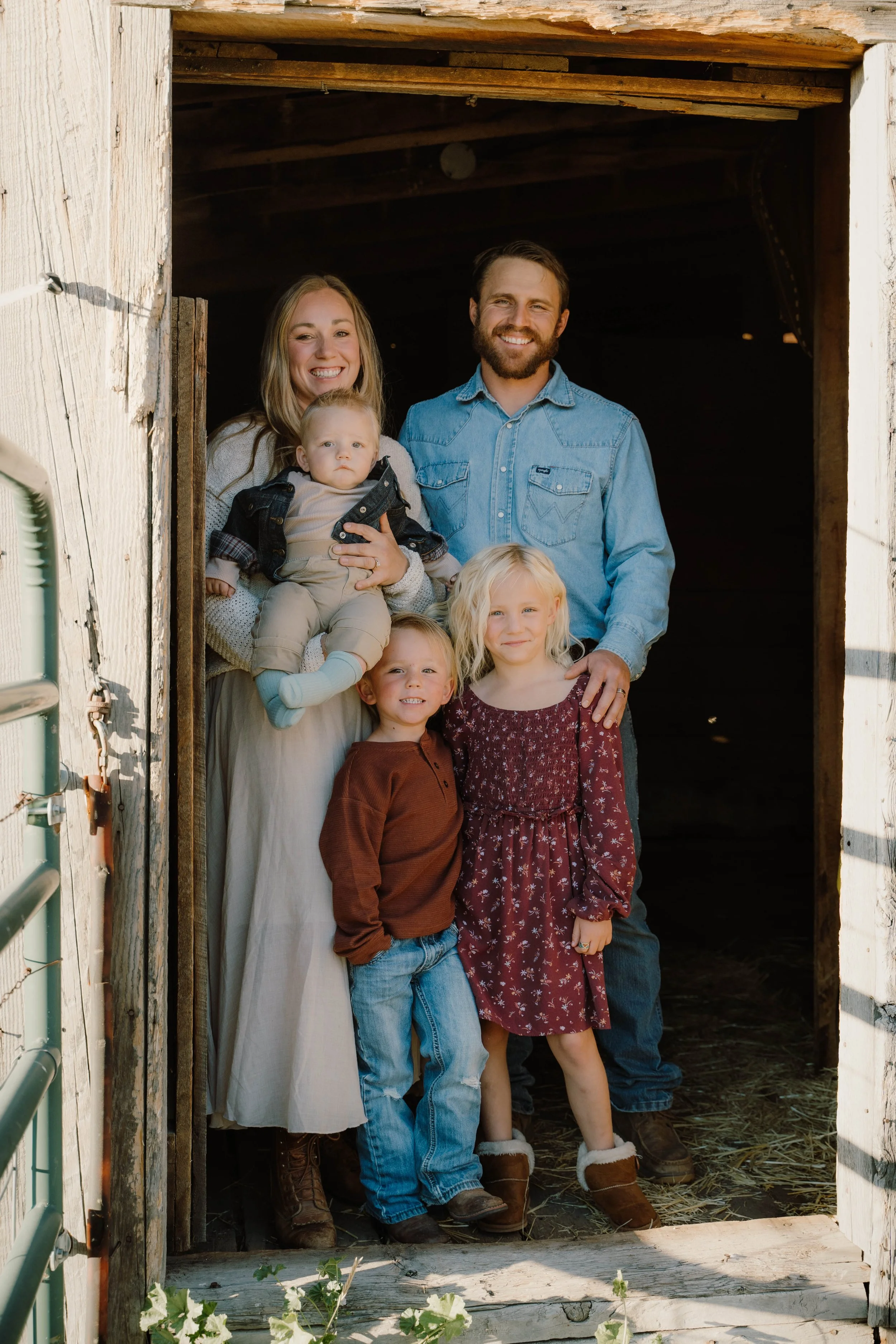 A family of six standing in a barn doorway, smiling at the camera. The family includes a woman, a man, and four young children. The barn has weathered wood and is partially open, with sunlight illuminating the group.