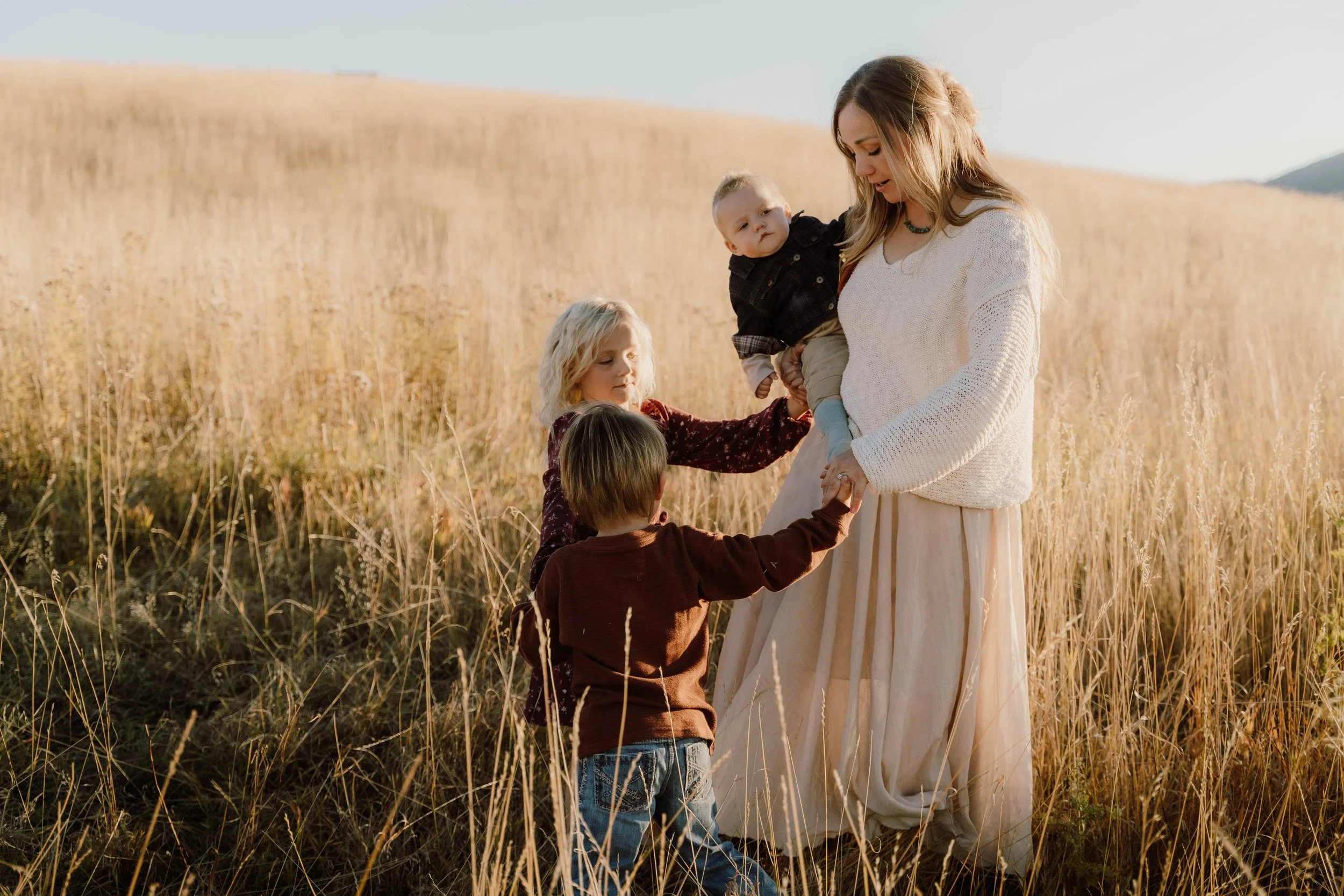 A woman with children in a golden wheat field during sunset, with mountains in the background.