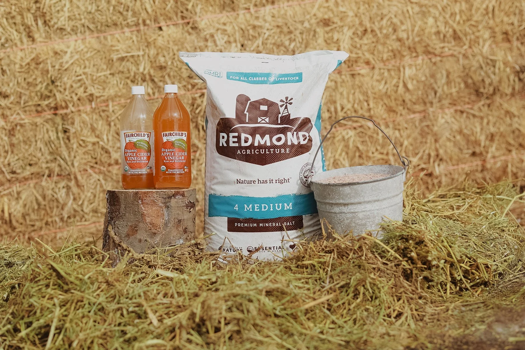 Bottled apple cider vinegar, a bag of Redmond mineral salt, and a metal bucket on a hay-filled surface.