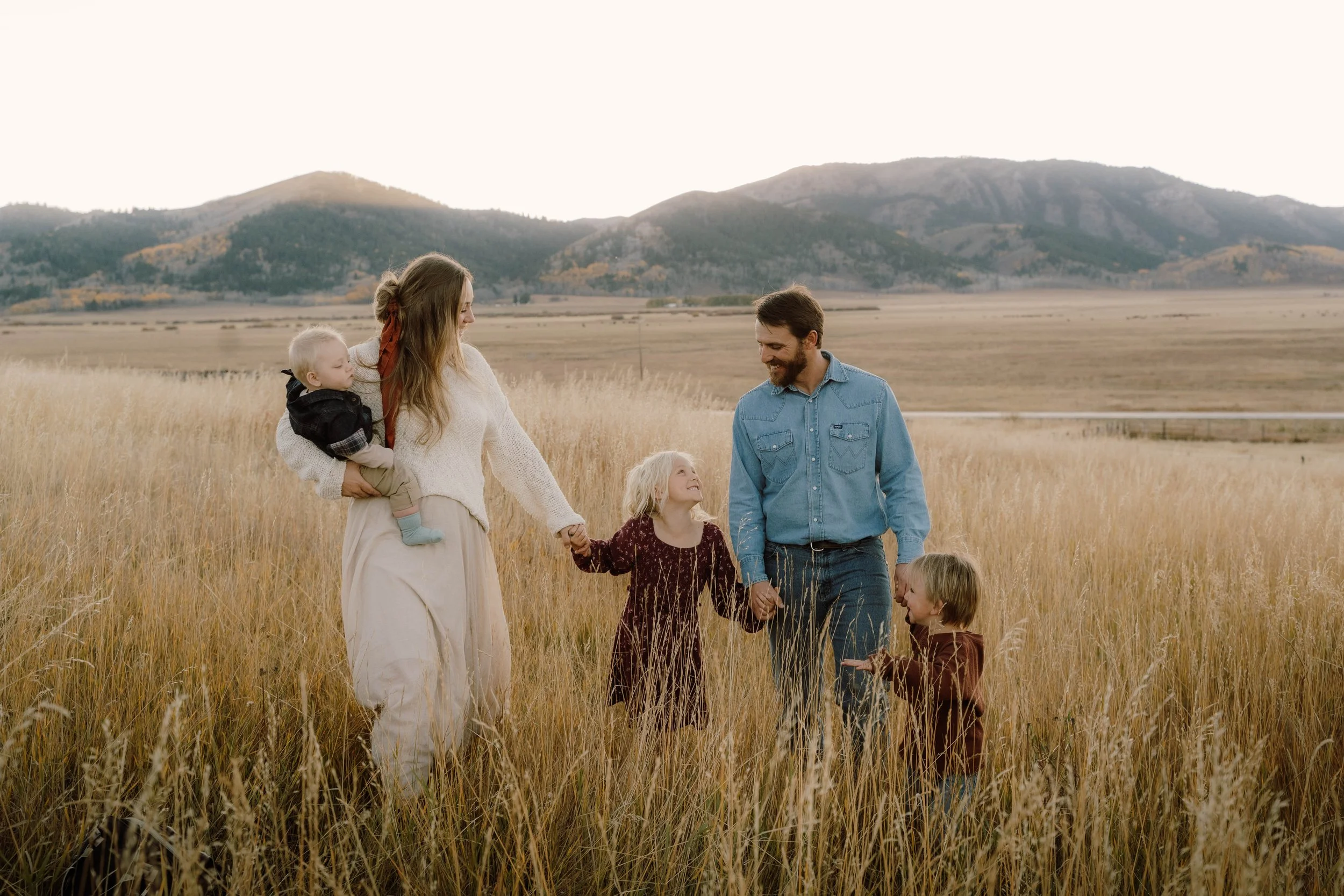 Family of five holding hands in a field of tall grass with mountains in the background during sunset.