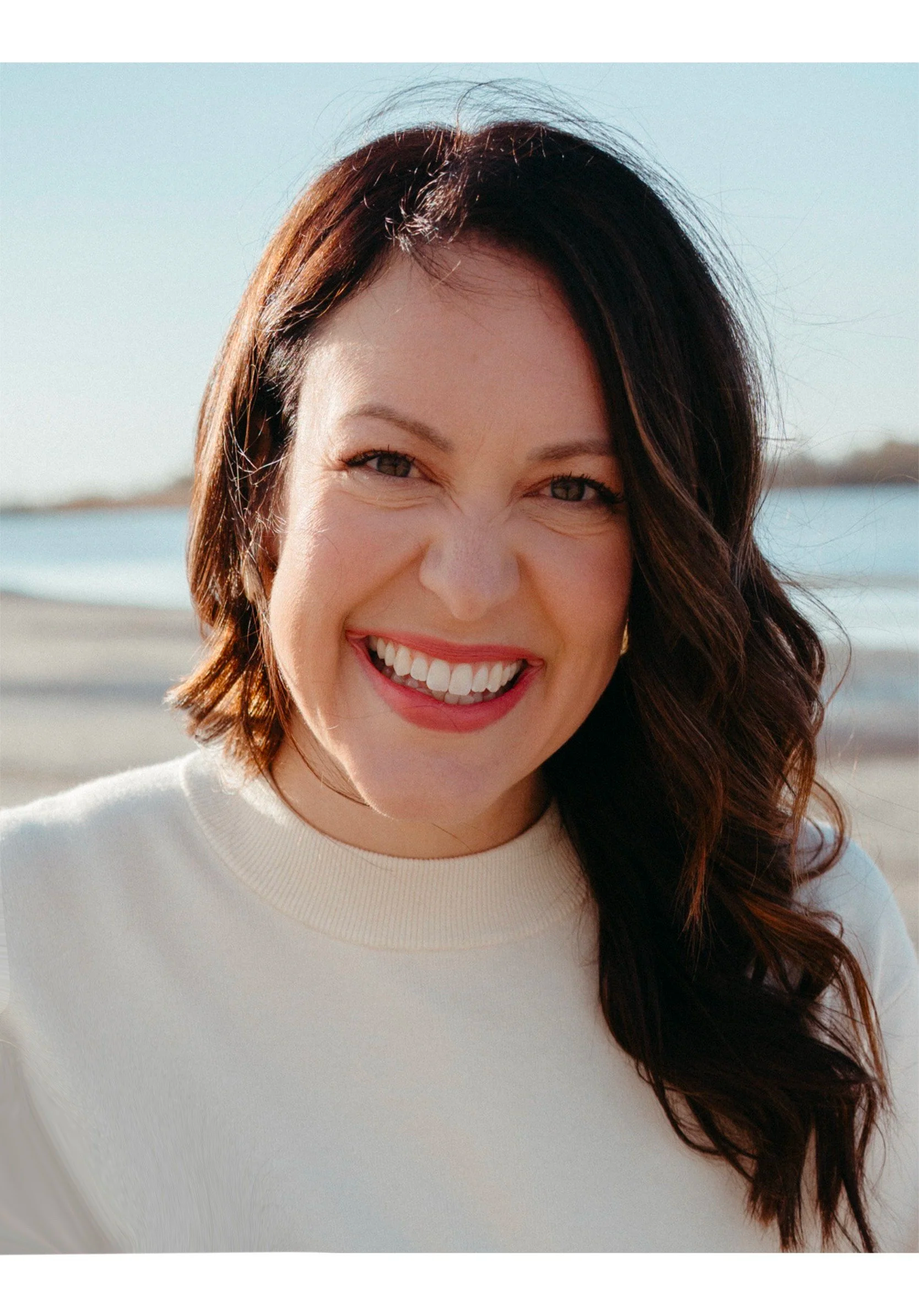 A smiling woman with brown hair and wearing a white top, standing outdoors near water, with a shoreline in the background.
