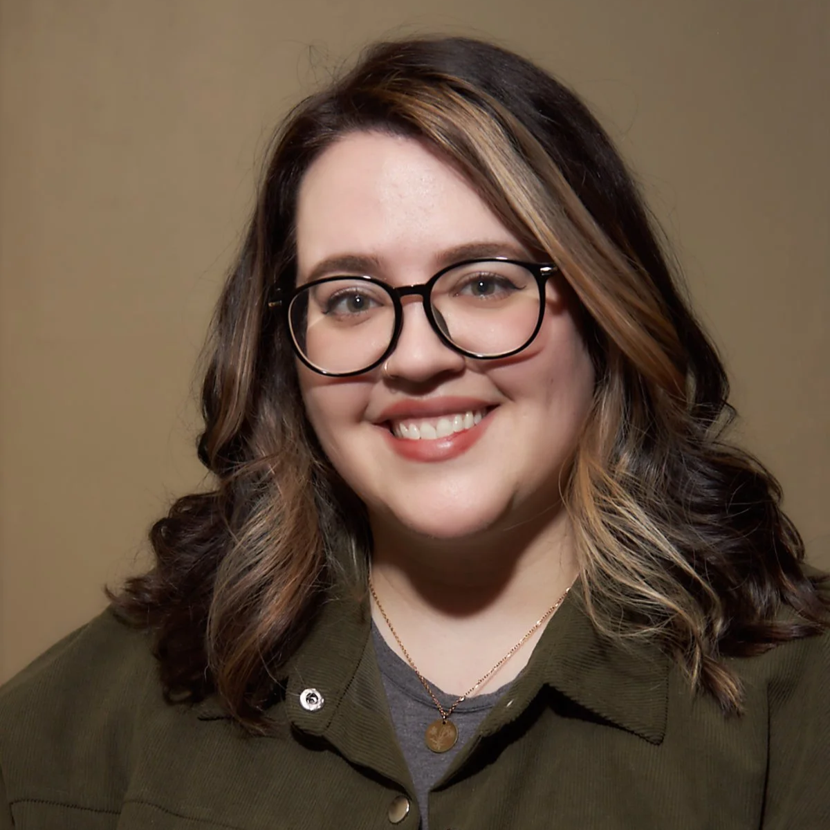 A young woman with glasses and shoulder-length wavy hair, smiling, wearing a green jacket and a necklace, against a plain background.