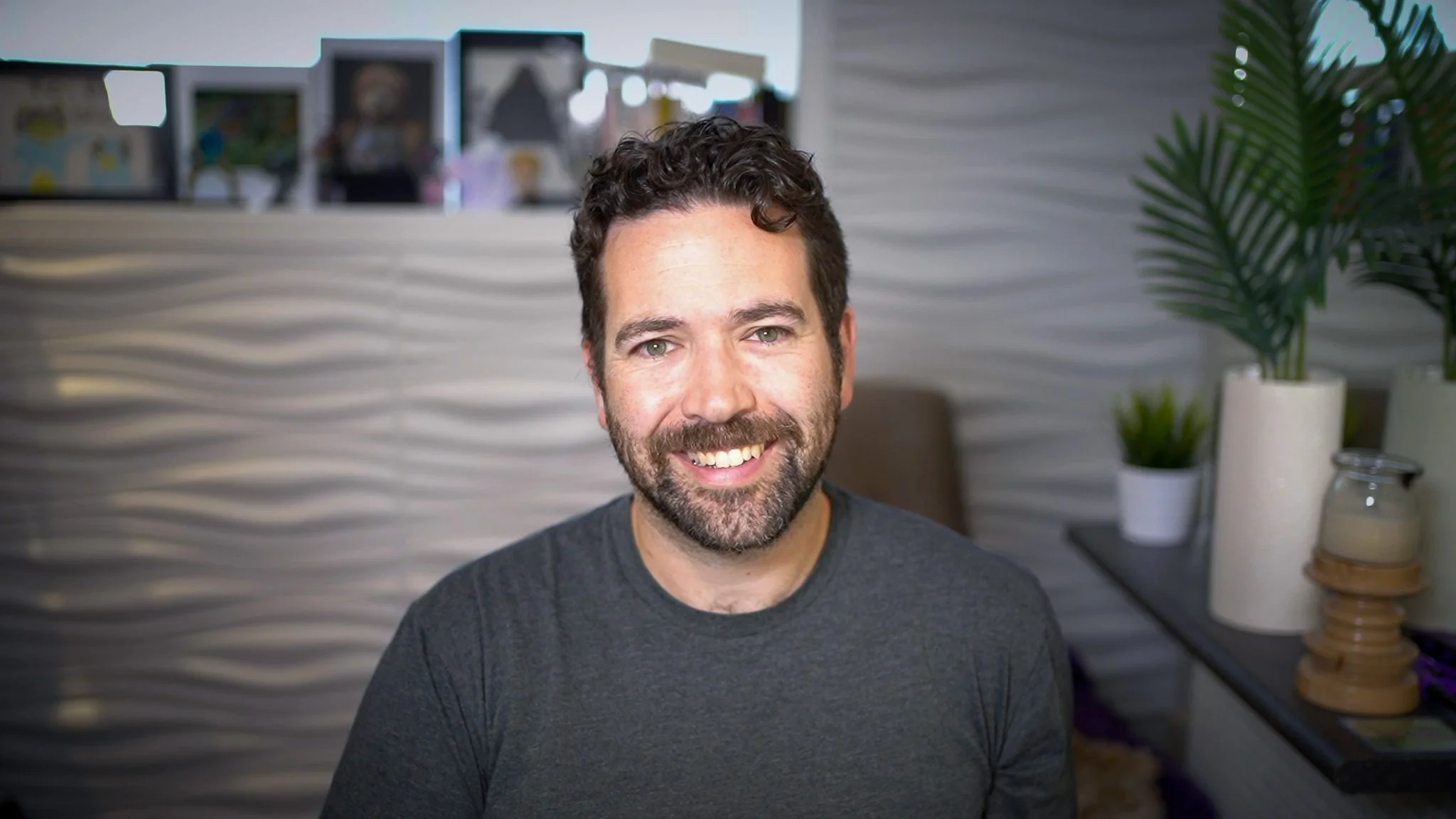 A smiling man with curly dark hair and a beard, wearing a dark gray t-shirt, sitting indoors in front of a textured white wall with potted plants and framed pictures in the background.