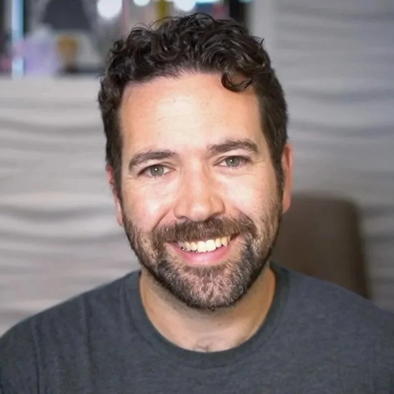 A smiling man with curly dark hair, a beard, and wearing a dark gray shirt, in front of a blurred background.