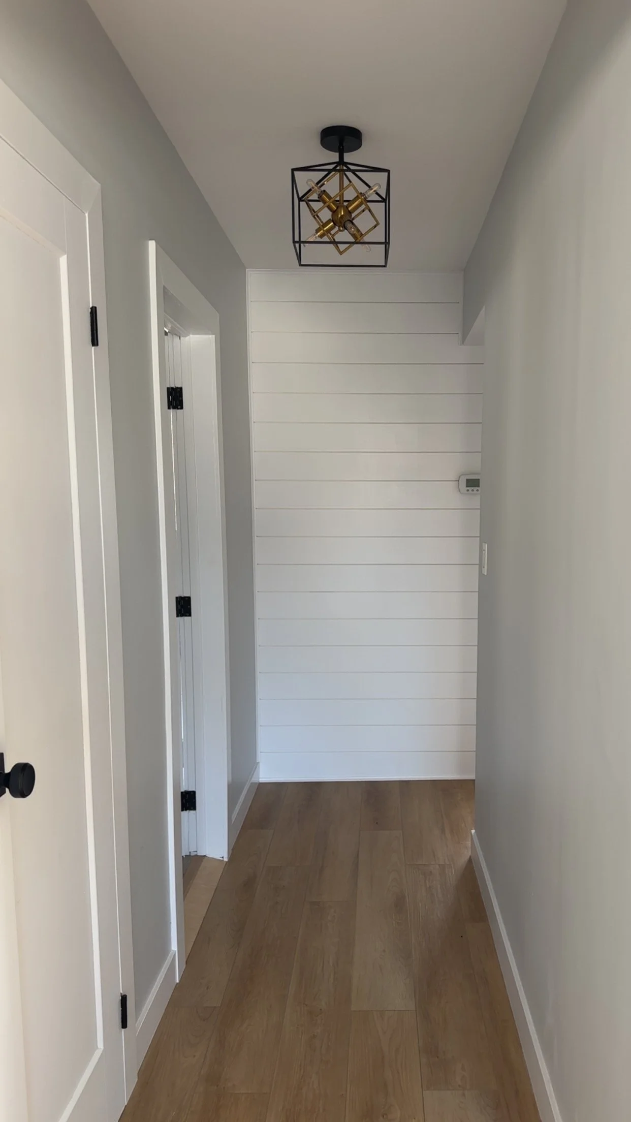 A narrow hallway with wood flooring. White shiplap accent wall at the end of the hallway with a digital thermostat. A modern black and gold geometric ceiling light fixture is installed on the ceiling. Azure Interiors Southwestern Ontario.