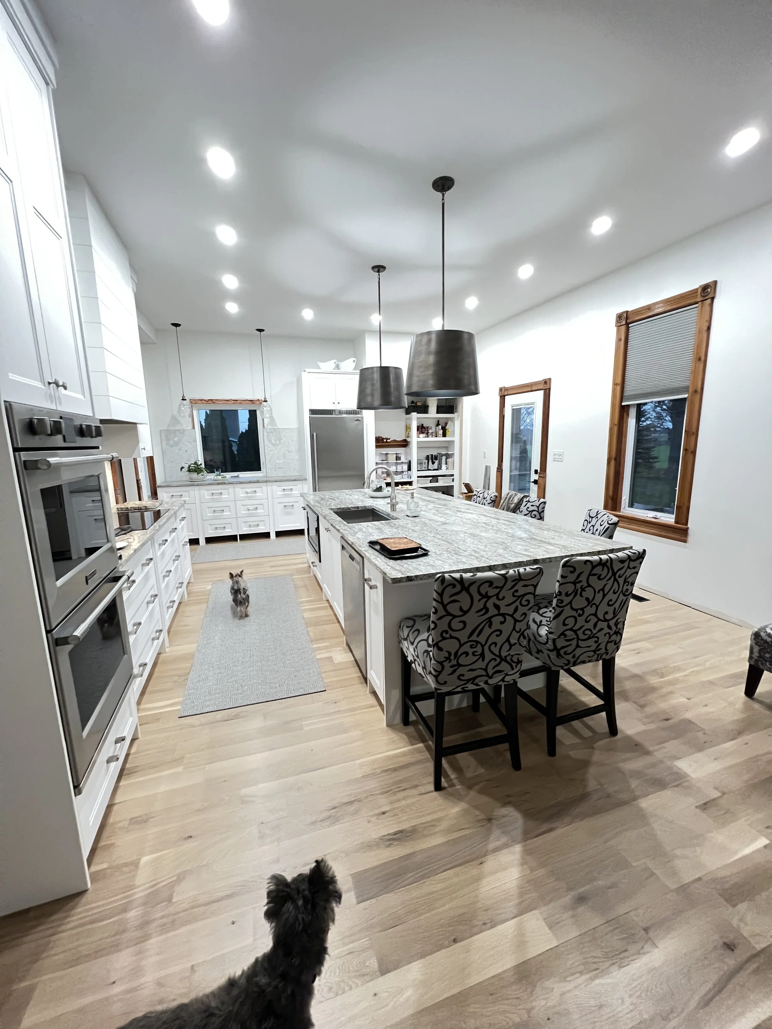 A wider look at the white kitchen, showing the long island, wall ovens, warm wood floors and space between the work areas.