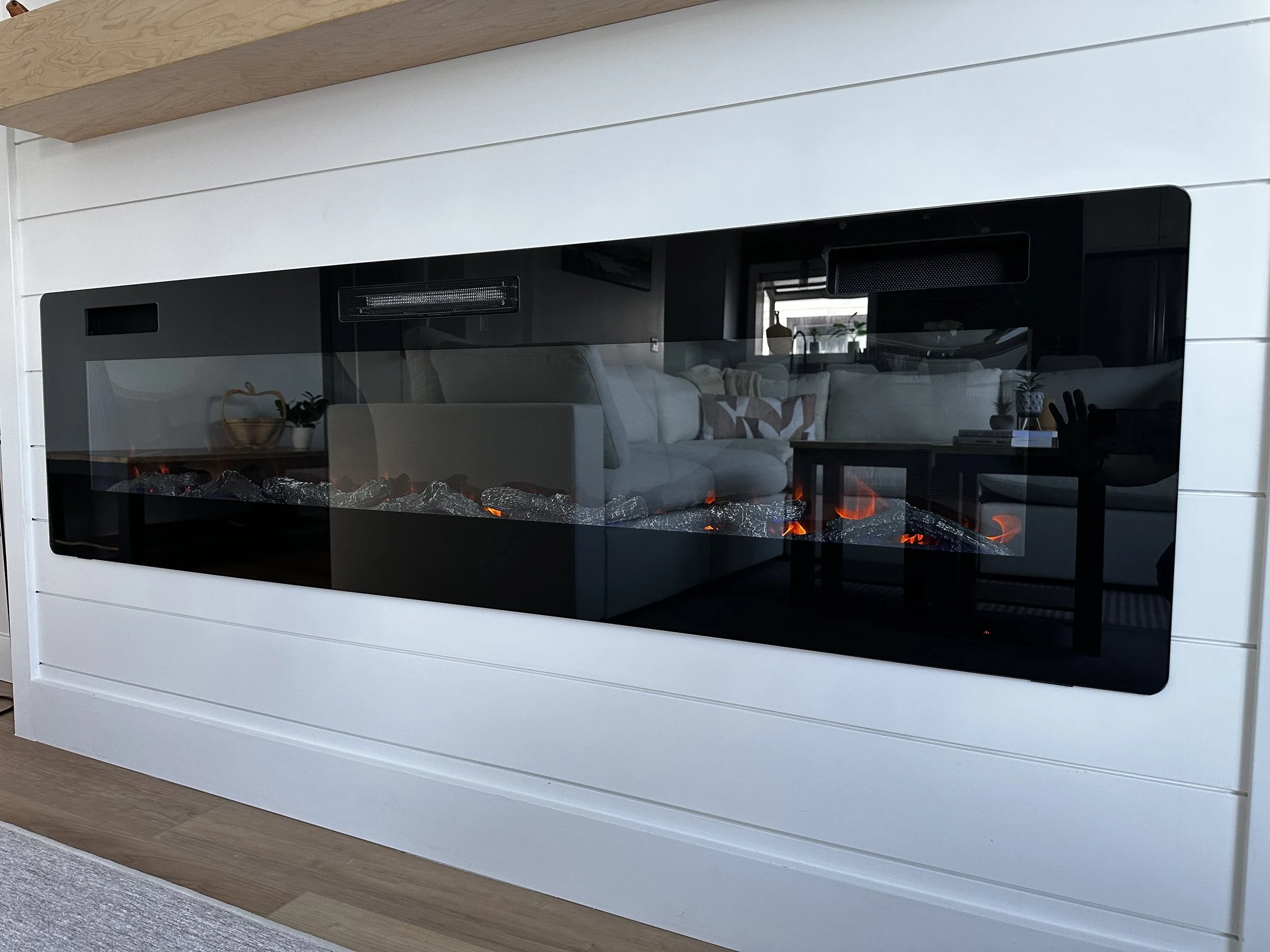Modern electric fireplace with a black glass front, built into a white paneled wall beneath a wooden shelf in a living room. Azure Interiors Southwestern Ontario.
