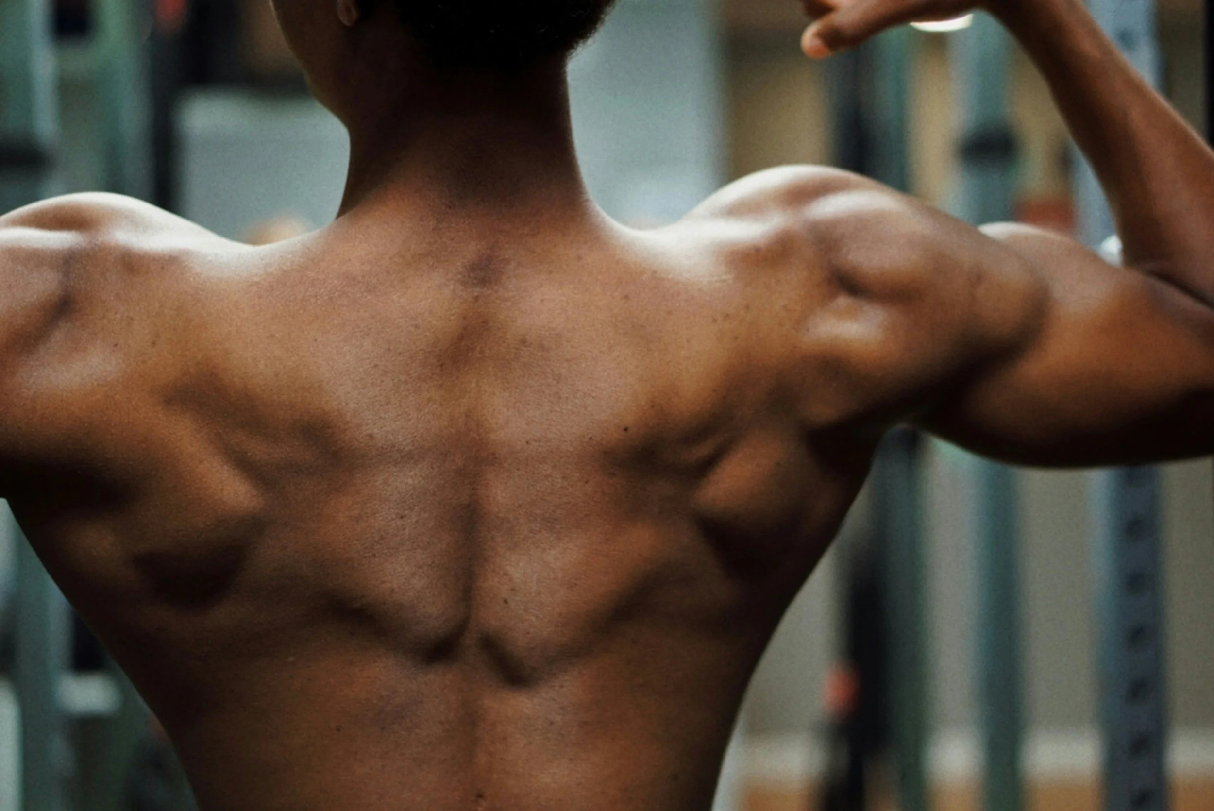 Close-up of a shirtless person with dark skin showing muscular back and shoulders, with one hand raised near the head, in what appears to be a gym or fitness setting.
