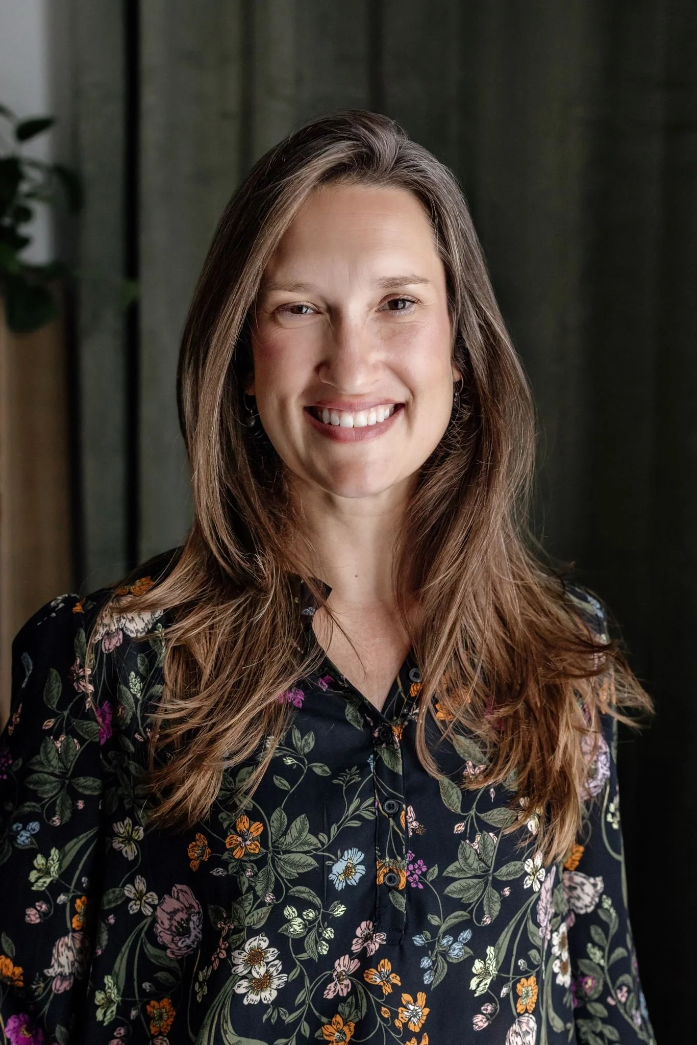 A woman with long, wavy hair smiling at the camera. She is wearing a black floral shirt with colorful flowers and leaves. The background appears to be indoors with dark curtains and a plant on the side.