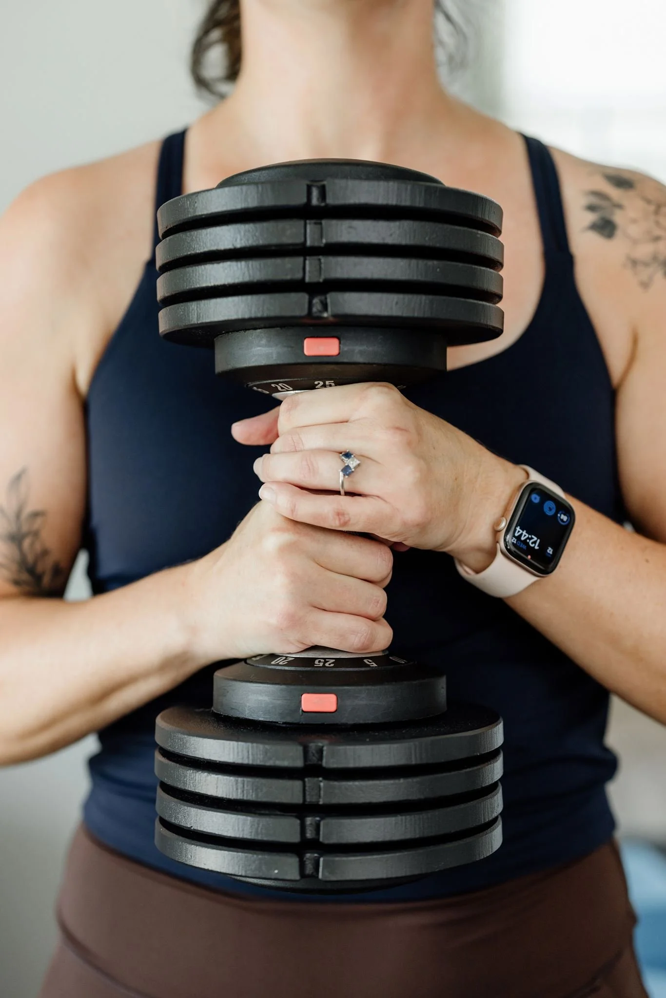 A woman wearing a sleeveless black top and a smartwatch holding a large adjustable black weight with multiple plates stacked in a vertical position.