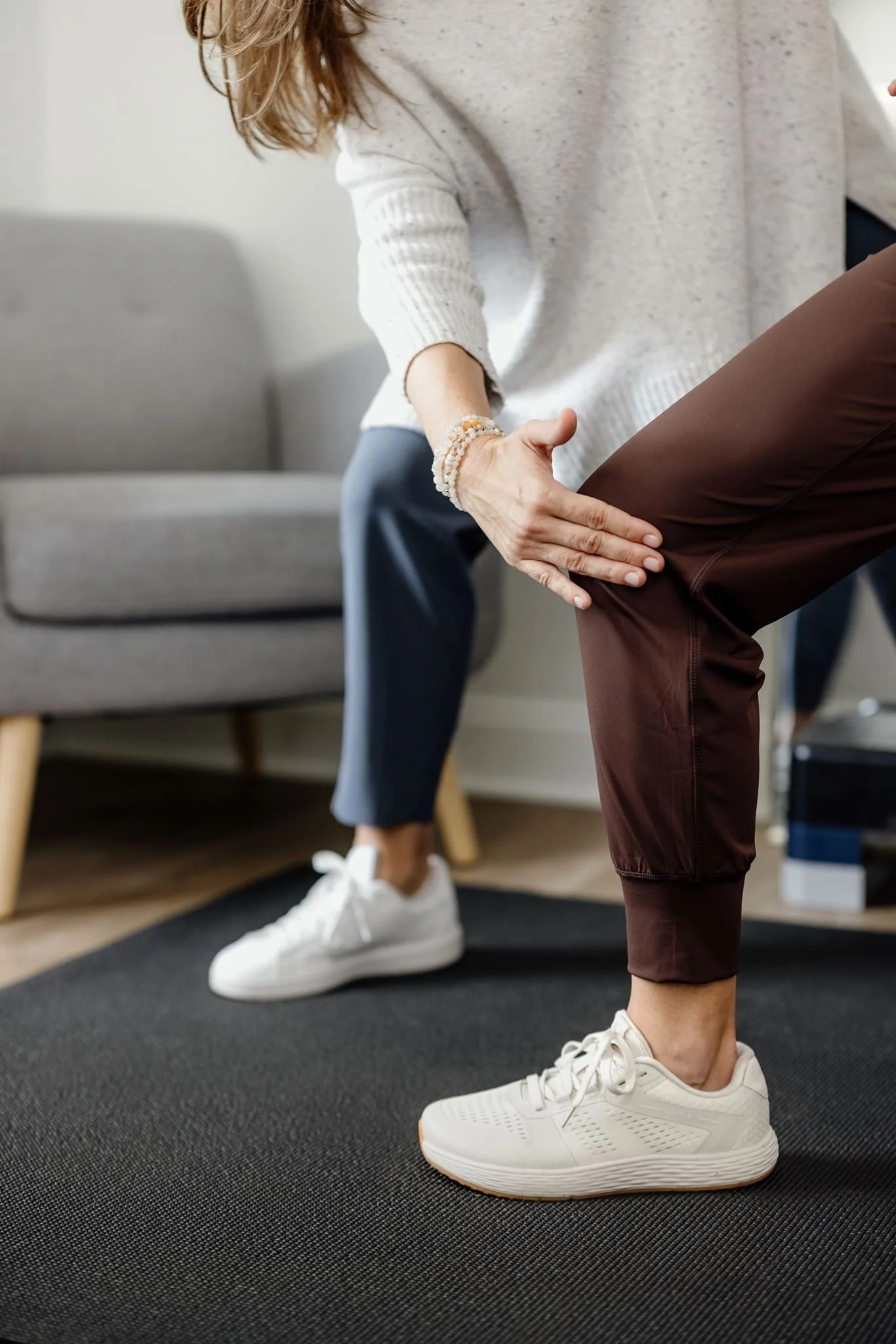 A person wearing white sneakers and brown pants standing on a dark exercise mat, with another person in the background wearing white sneakers, gray pants, and a white sweater, in a room with a gray sofa.