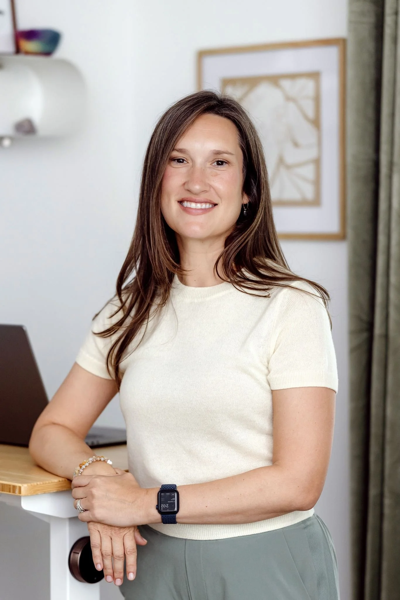 A woman with long brown hair smiling while standing with her arms crossed inside a room with framed artwork on the wall and a white shelf in the background.