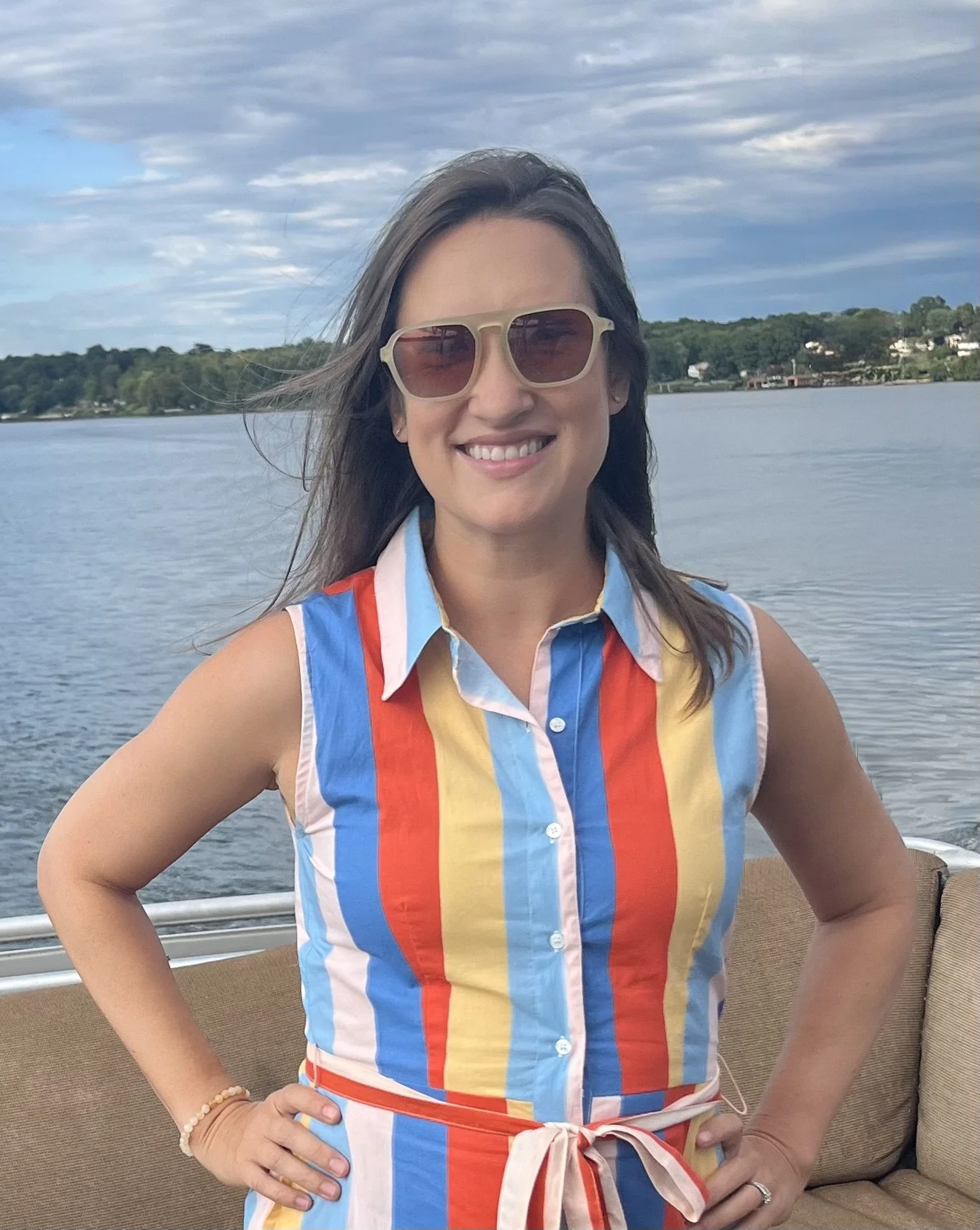 Smiling woman wearing striped sleeveless shirt and sunglasses, standing near water with trees and buildings in the background.