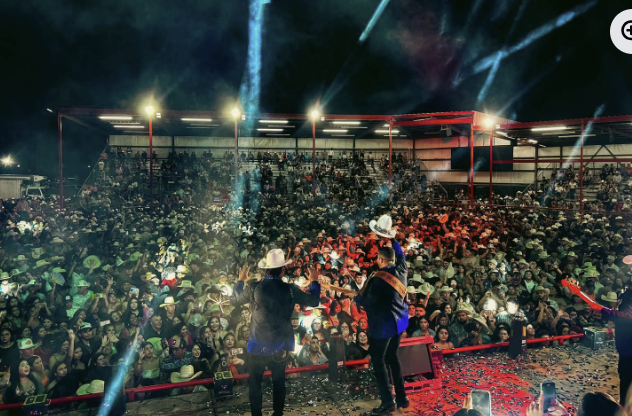 A concert at a rodeo arena with a large crowd, two musicians in cowboy hats singing and playing guitar on stage, colorful lighting, and confetti on the ground.