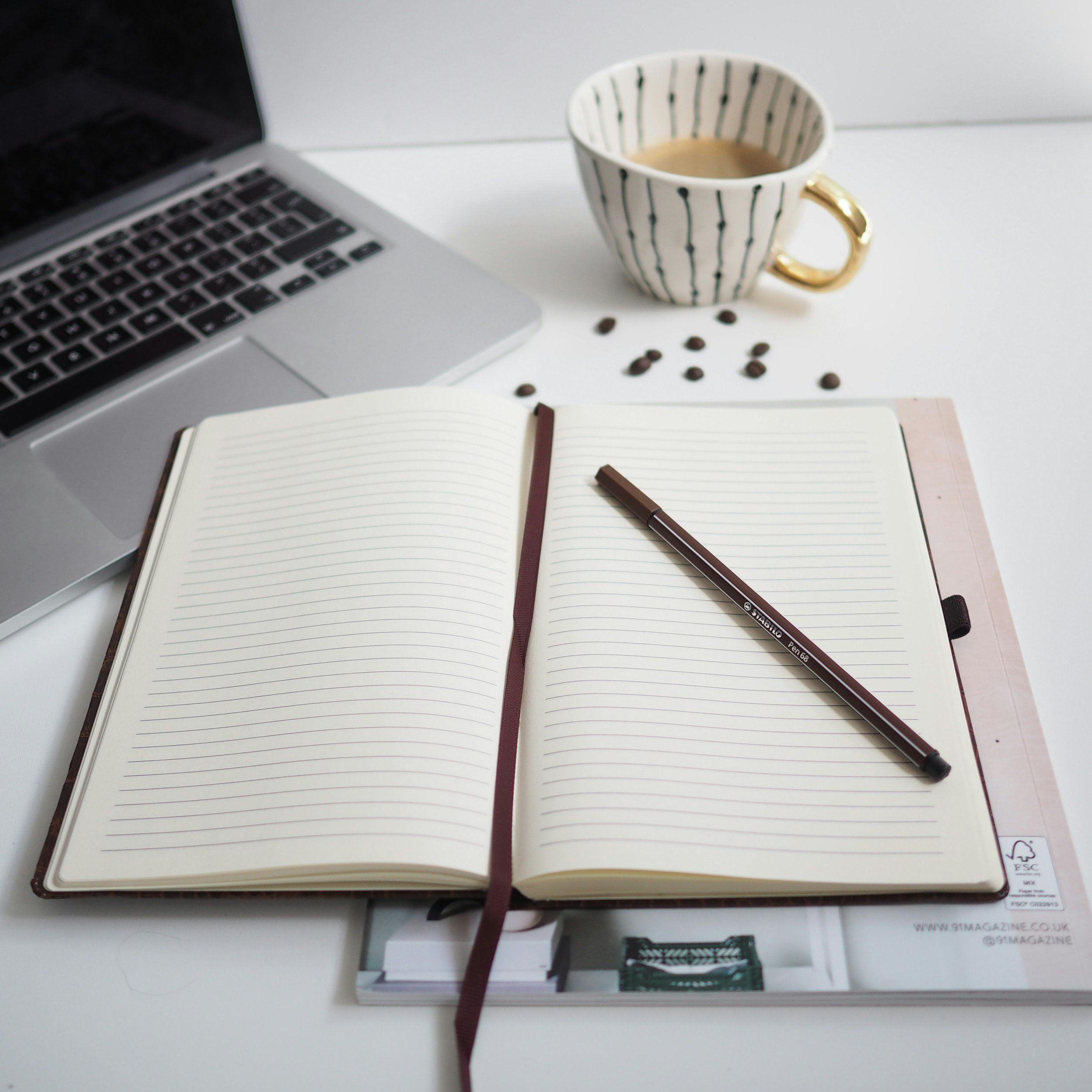 Open notebook with lined pages, a brown pen, spilled coffee beans, a striped coffee cup with coffee, a laptop, part of a printed magazine, and a card on a white surface.