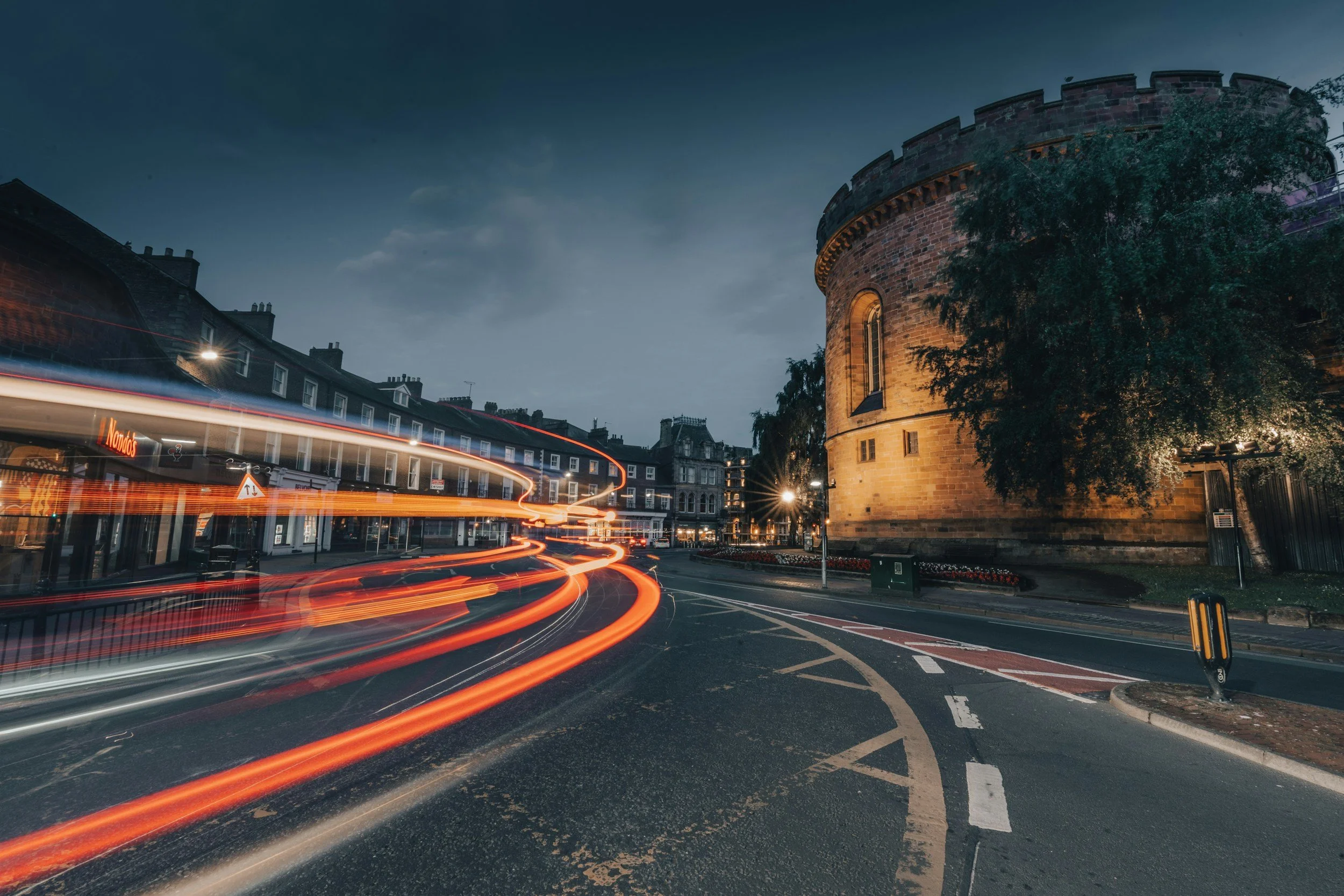 Historic stone building with a rounded tower, trees, and a street at dusk, with light trails from moving vehicles in the foreground.