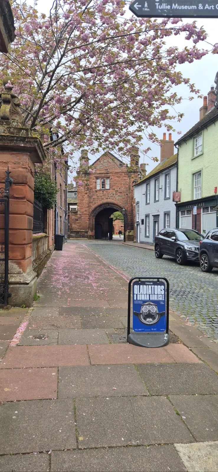 A cobblestone street with pastel-colored buildings, a cherry blossom tree with pink flowers, and a historic arched gateway made of red brick in the background. There is a signboard in the foreground advertising a Roman Carlisle Gladiators show.