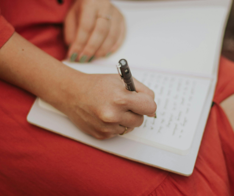 A person writing in a notebook with a black pen, sitting on a red surface.