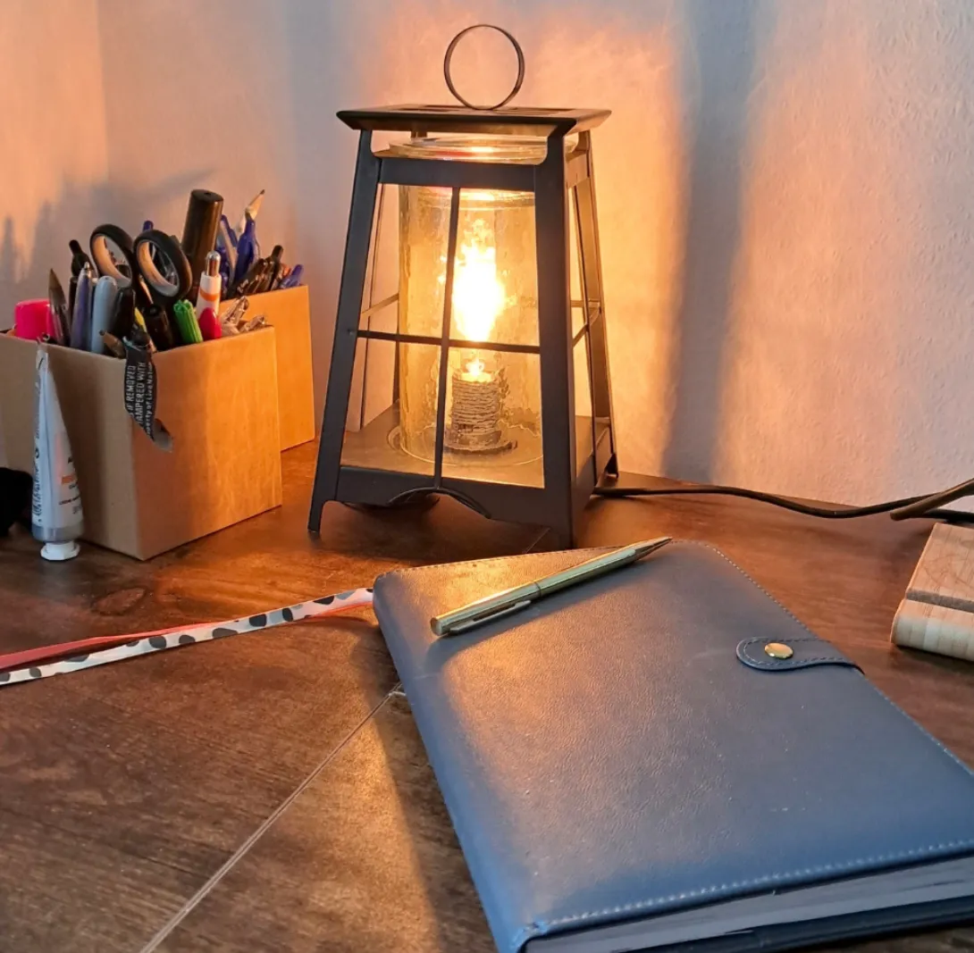 A desk with a blue notebook and a pen, a film lantern emitting warm light, a container of various office supplies, and a ruler with a striped pattern.