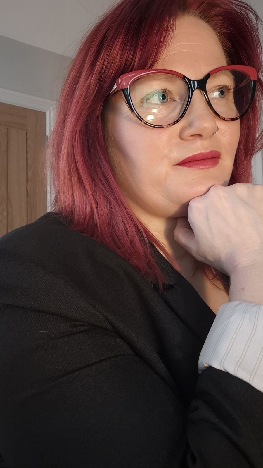 A woman with red hair and glasses, wearing red lipstick, posing with her chin resting on her hand.