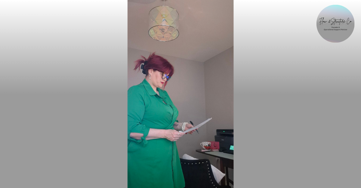 A woman with red hair and glasses standing in a room, reading a document.