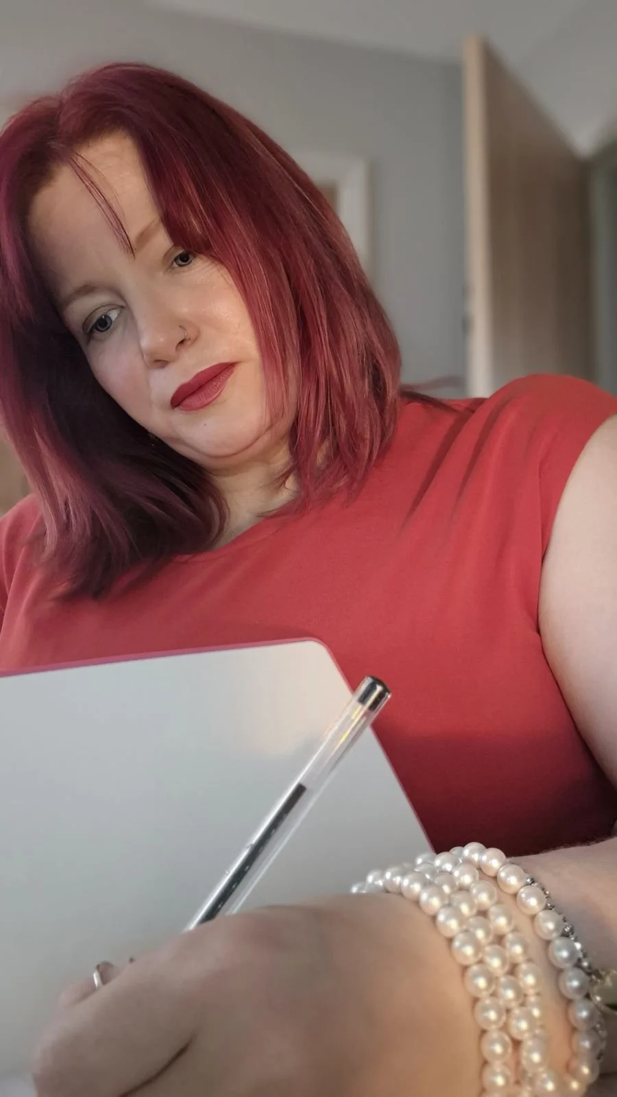 A woman with red hair and wearing a red top is holding a white notebook and a pen. She is also wearing pearl bracelets on her wrist.