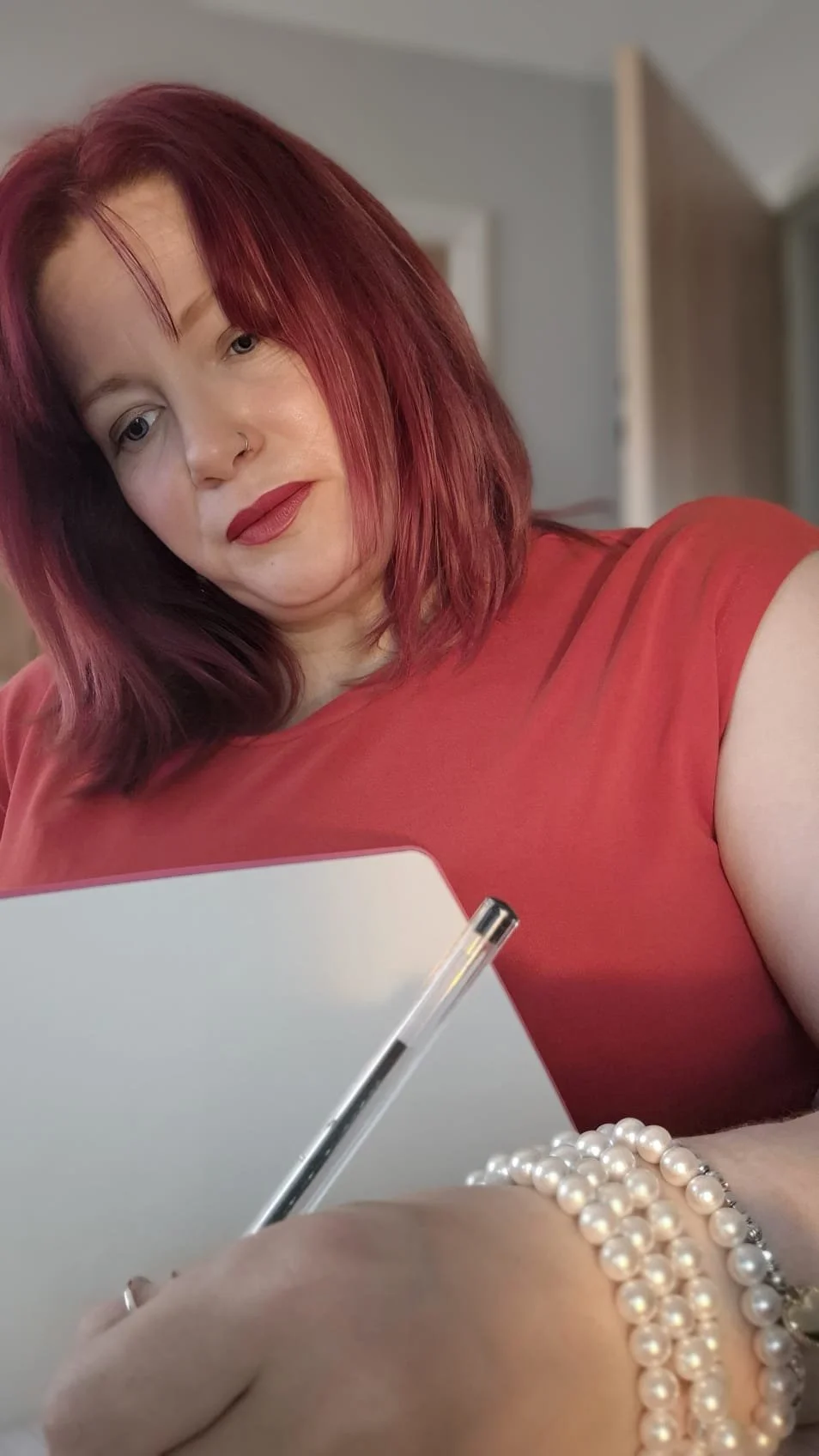 A woman with red hair wearing a red top and pearl bracelets, holding a white notebook and pen, looking at the camera.