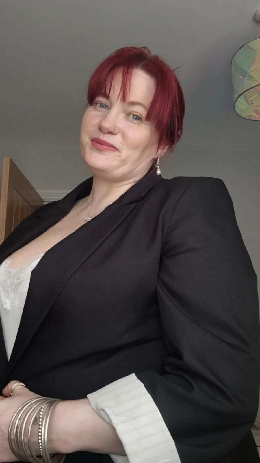 A woman with short red hair, wearing a black blazer, light-colored blouse, and jewelry, poses for a photo indoors with a gray ceiling and colorful lamp in background.