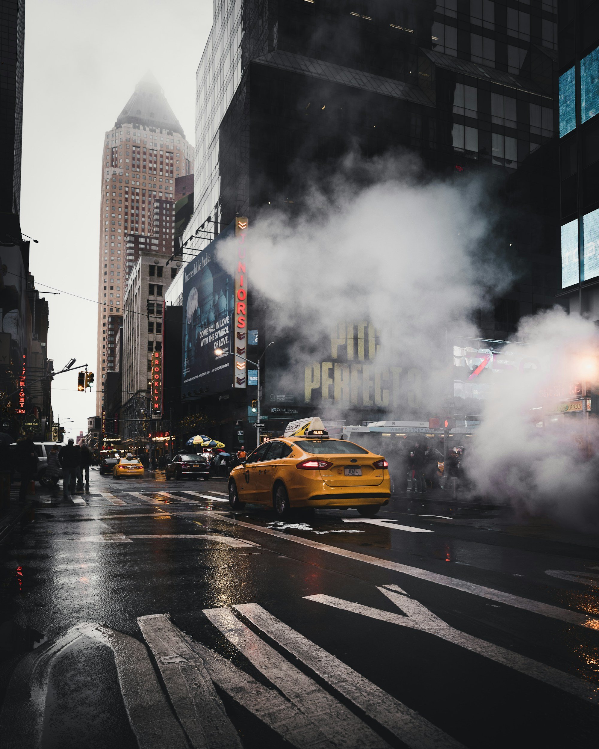 A city street in Manhattan during rainy weather with black asphalt, yellow taxis, pedestrians, and foggy atmosphere, and tall buildings with digital billboards.