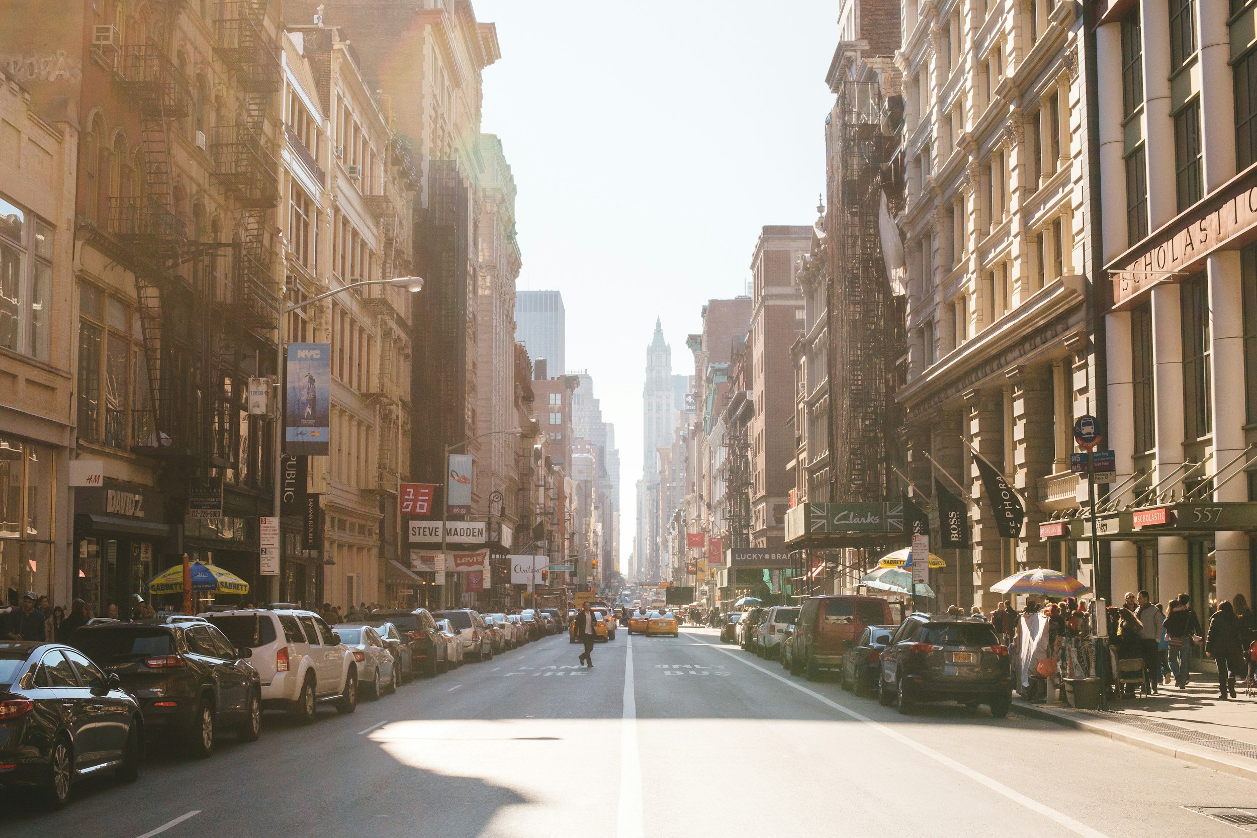 A busy city street with tall buildings on both sides, parked cars along the curb, people walking on the sidewalks, and various signs for stores and brands hanging from the buildings, with a bright sky overhead.