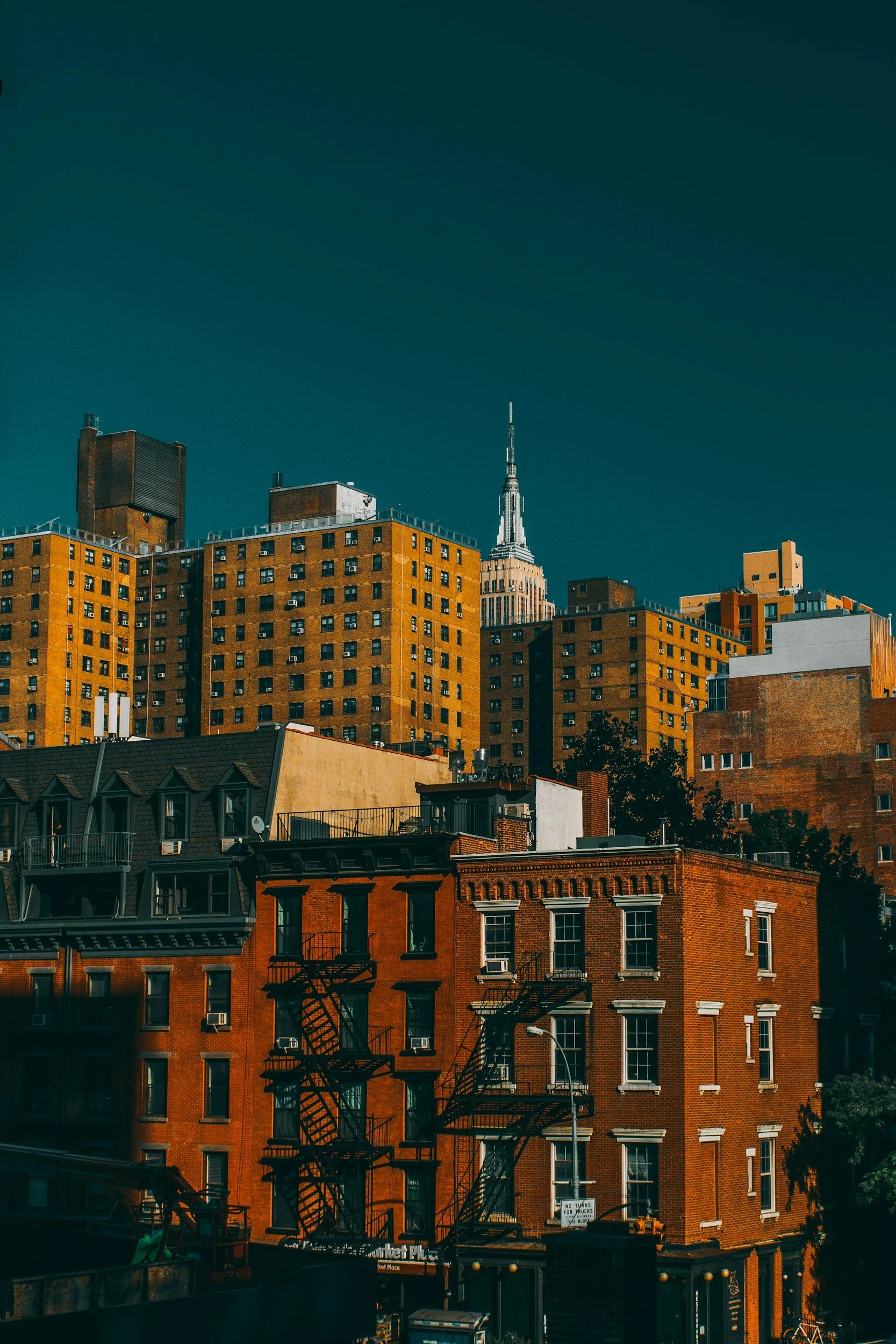 City skyline with tall buildings, including the Empire State Building, seen against a clear sky with a gradient from dark blue to teal. The foreground shows older brick buildings with fire escapes and a street scene.