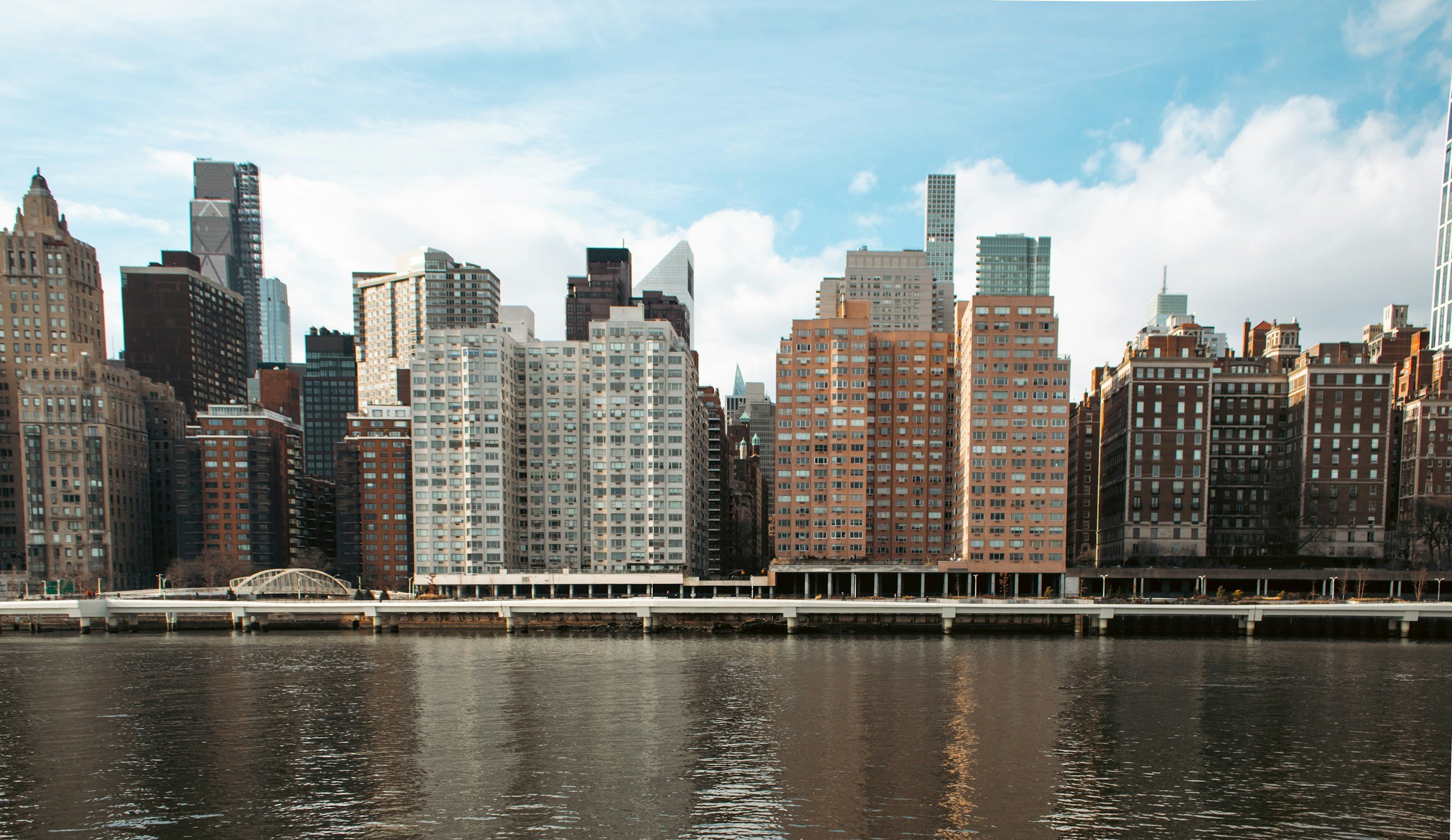 A city skyline with tall buildings viewed over a body of water, under a partly cloudy sky.