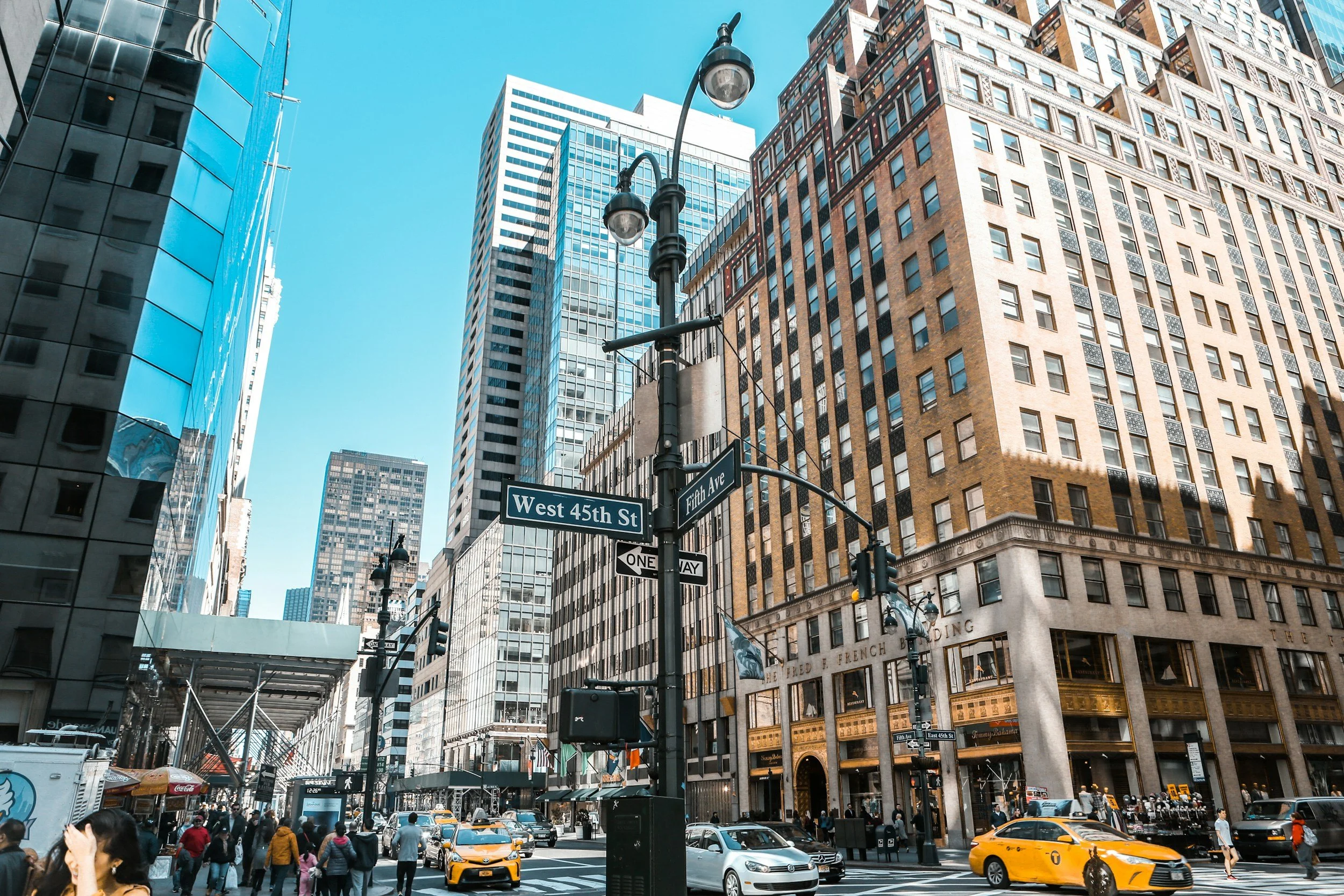 City street scene at the intersection of West 45th Street and Fifth Avenue in New York City with tall modern buildings, a street lamp, and traffic including yellow taxis and pedestrians walking.