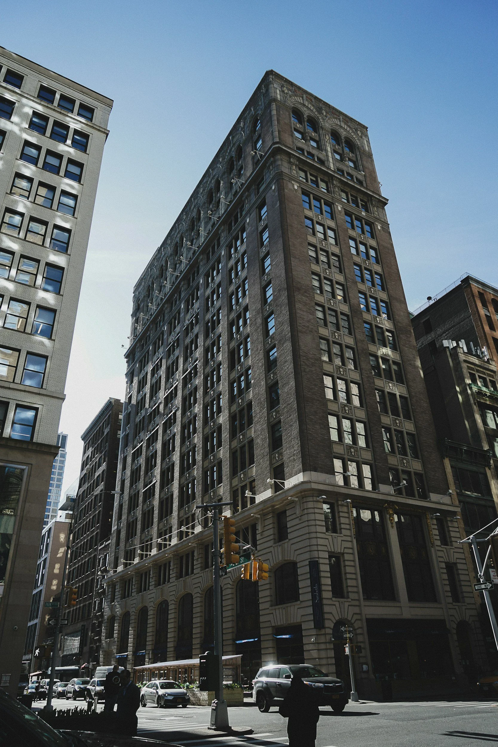 A city street scene with tall office buildings, some with reflective windows, cars driving and parked along the street, and pedestrians walking at the crosswalk. Traffic lights and street signs are visible.