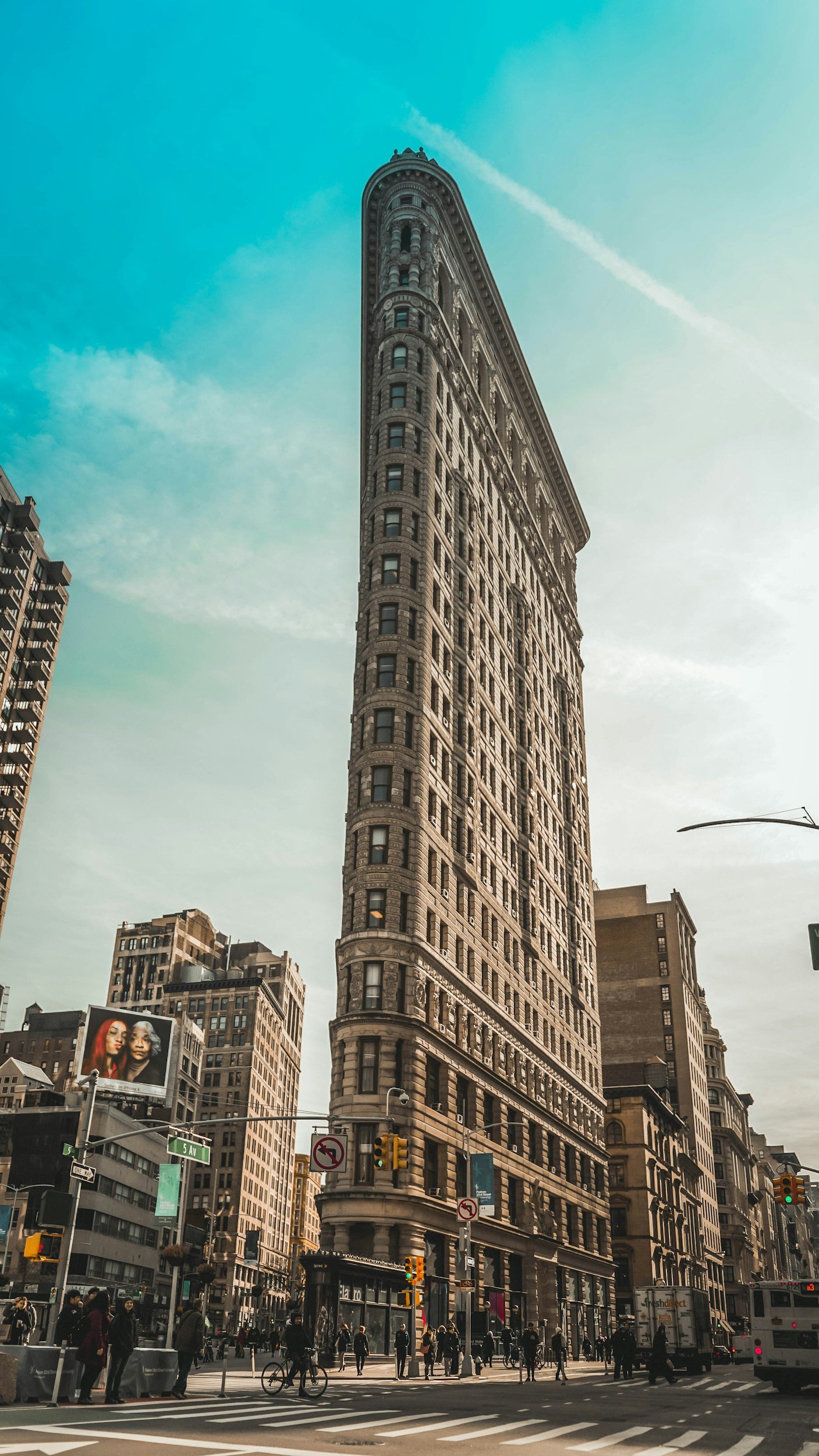 Famous flatiron building in New York City with people walking and traffic at street level, billboards, and surrounding buildings under a partly cloudy sky.