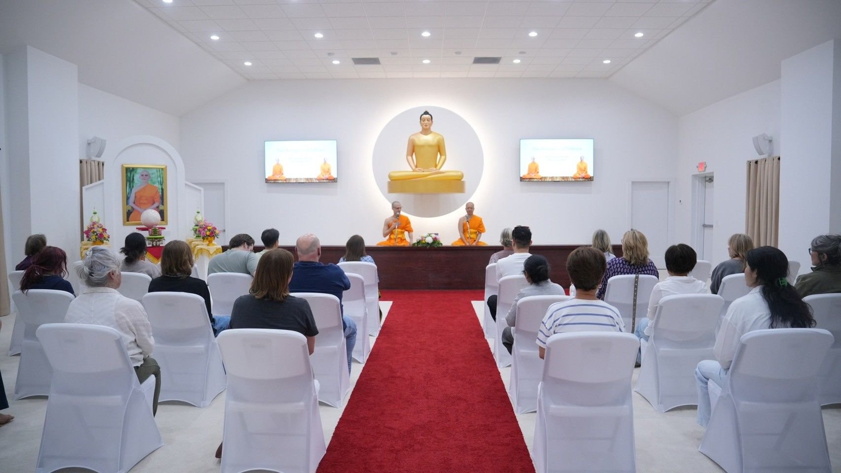 People seated in a room listening to two monks speaking at the front, with a large Buddha statue and smaller framed images of Buddha on the wall behind them.