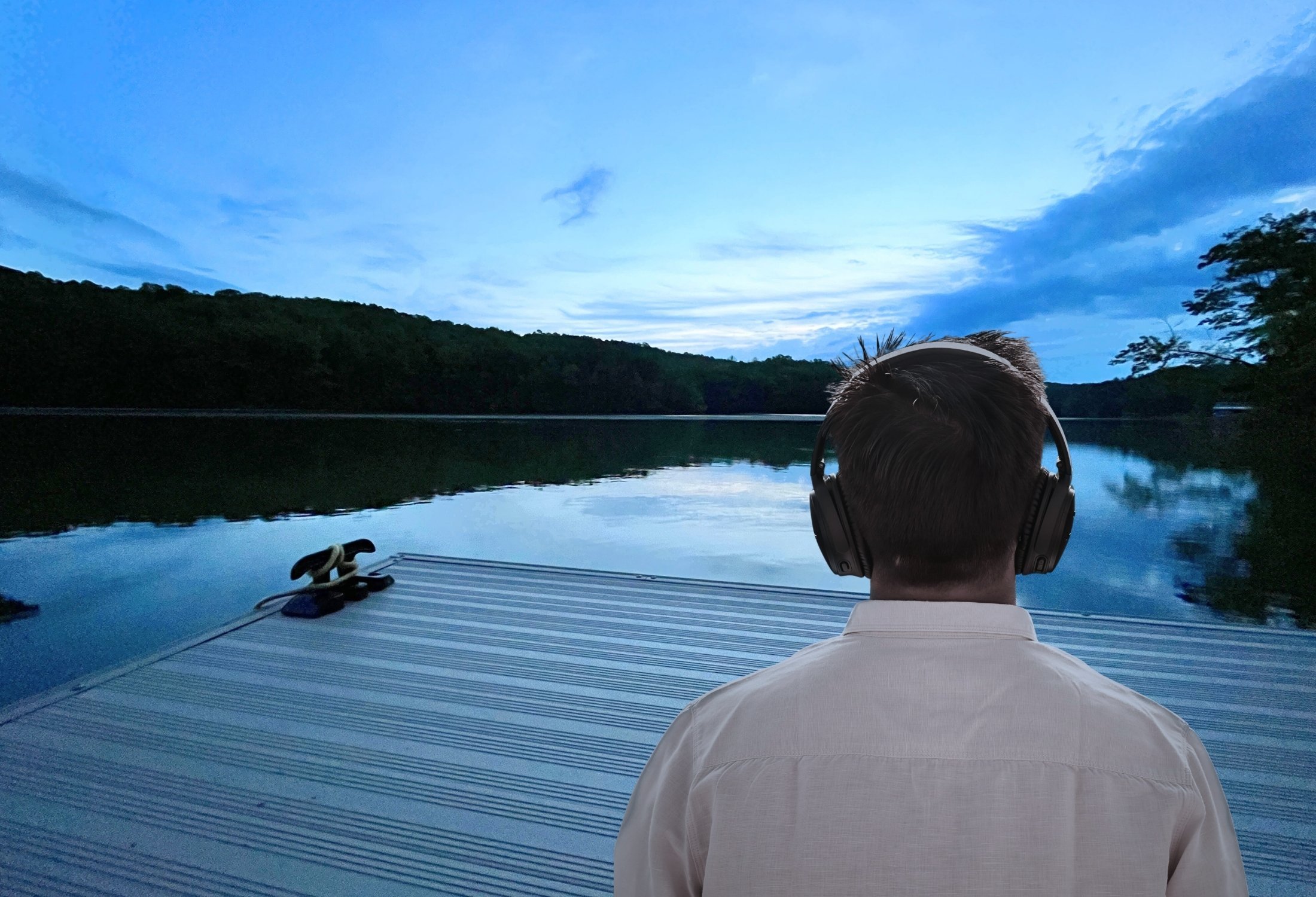A person with short hair is sitting on a boat, wearing headphones, facing a calm river with trees on both sides, under a partly cloudy sky in the evening or early morning.