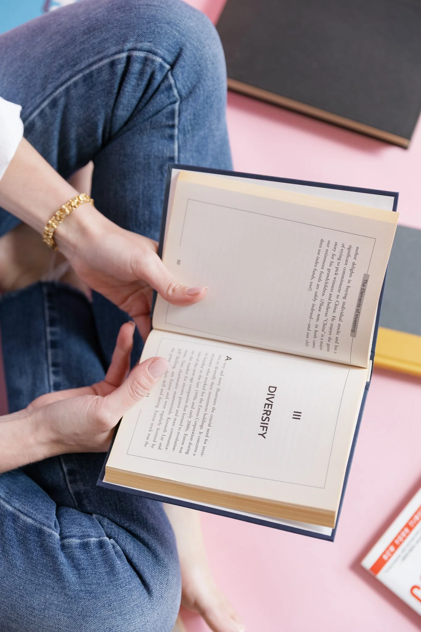Alex Merola sitting on the floor, wearing blue jeans, holding an open book titled 'DIVERSIFICATION' at a bookstore or library with pink flooring and bookshelves.