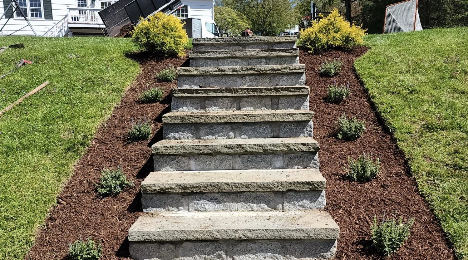 Outdoor stone staircase with a total of ten steps, flanked by small shrubbery plants and a mulched garden bed on both sides, leading up to a building with a small yard and other garden elements in the background.
