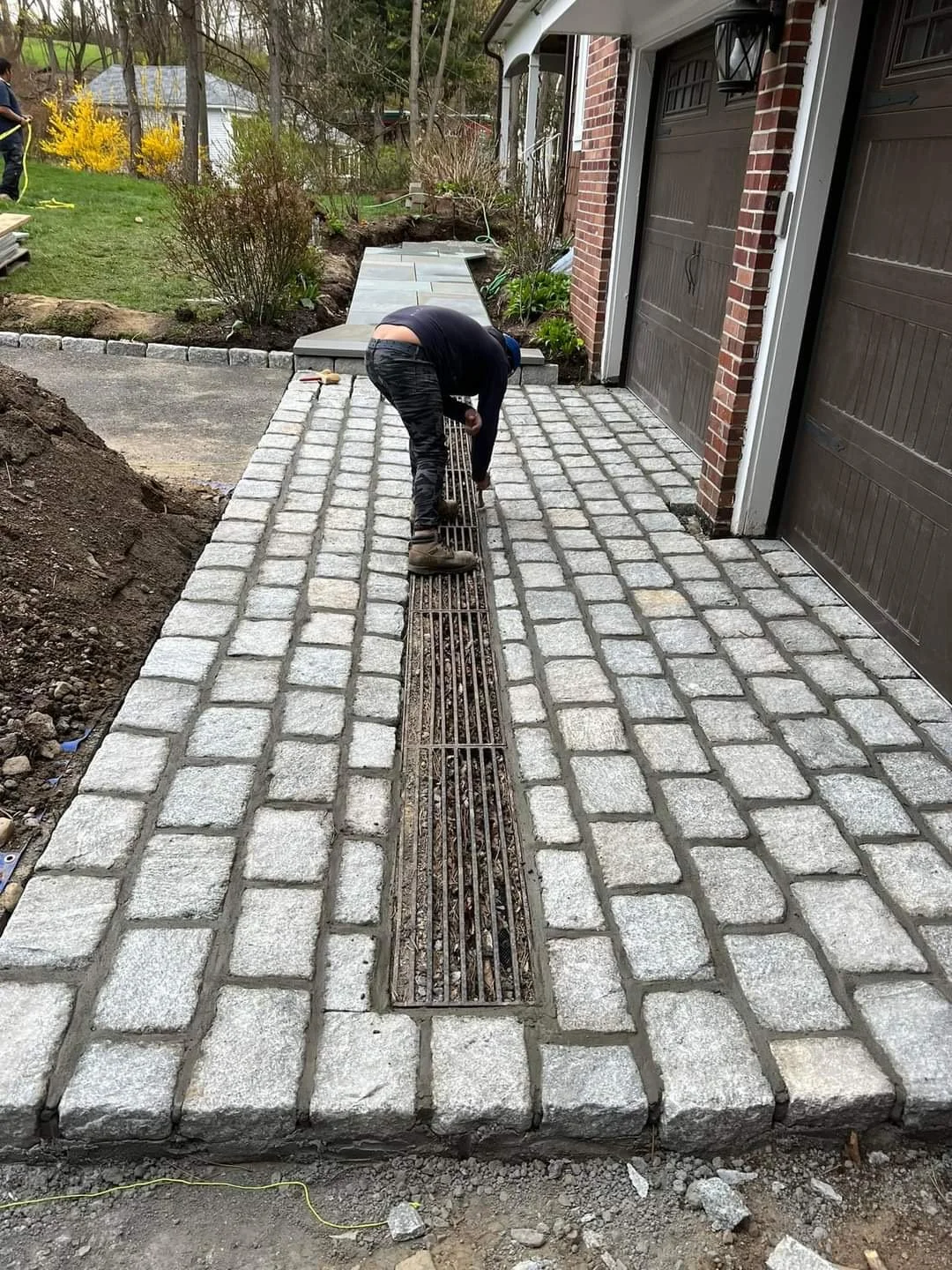 Person installing a brick border around a concrete driveway with a metal drain cover, next to a brick house with garage doors.