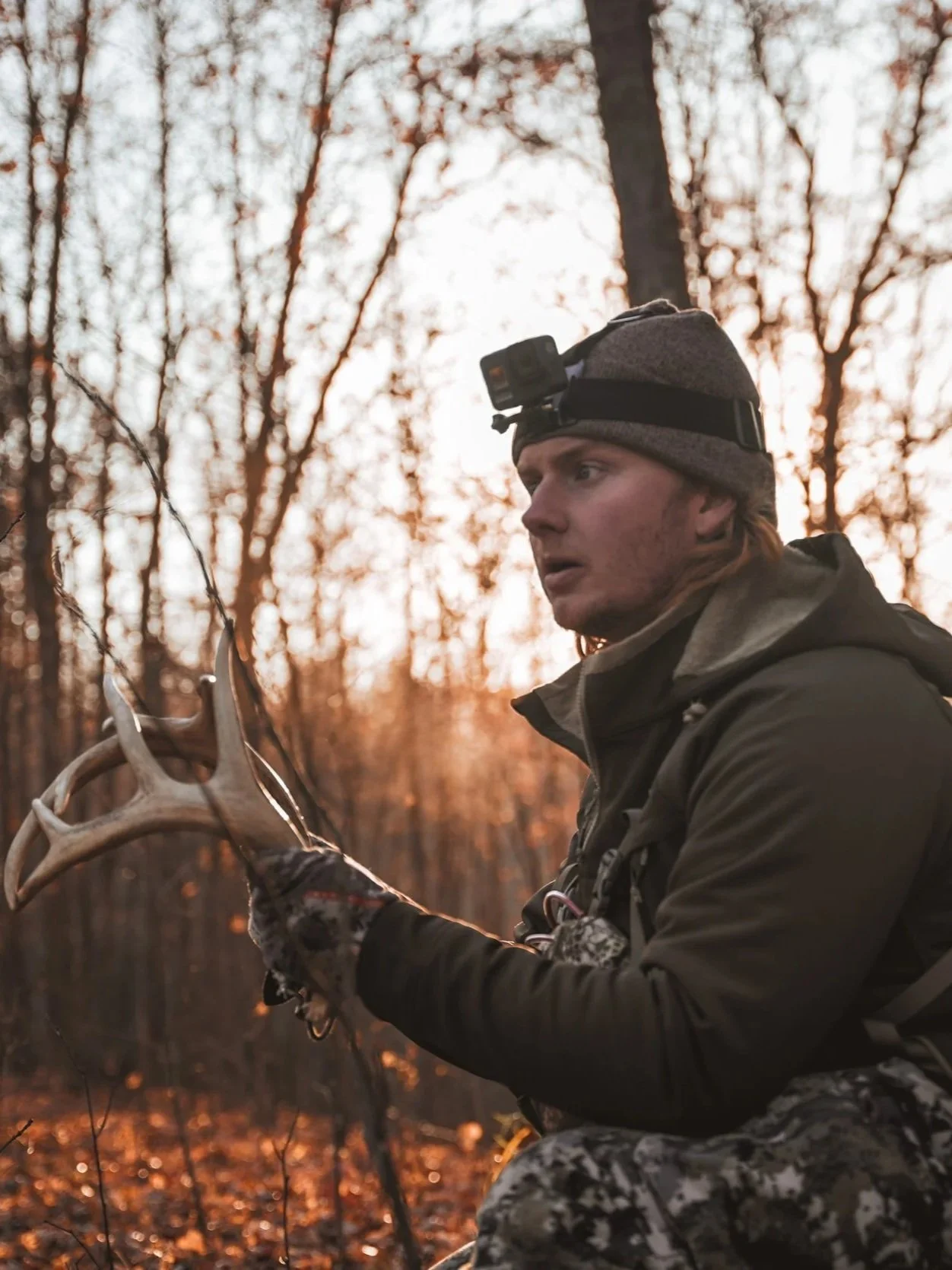 A man in outdoor hunting gear holding a shed antler in a forest during sunset, with trees and fallen leaves in the background.