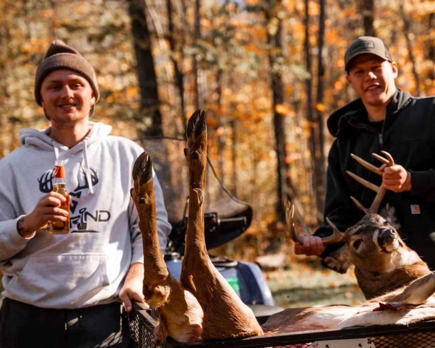 Two young men outdoors in a forest with autumn foliage, one holding a bottle and the other holding a deer skull and antlers, with a harvested buck on an ATV between them.