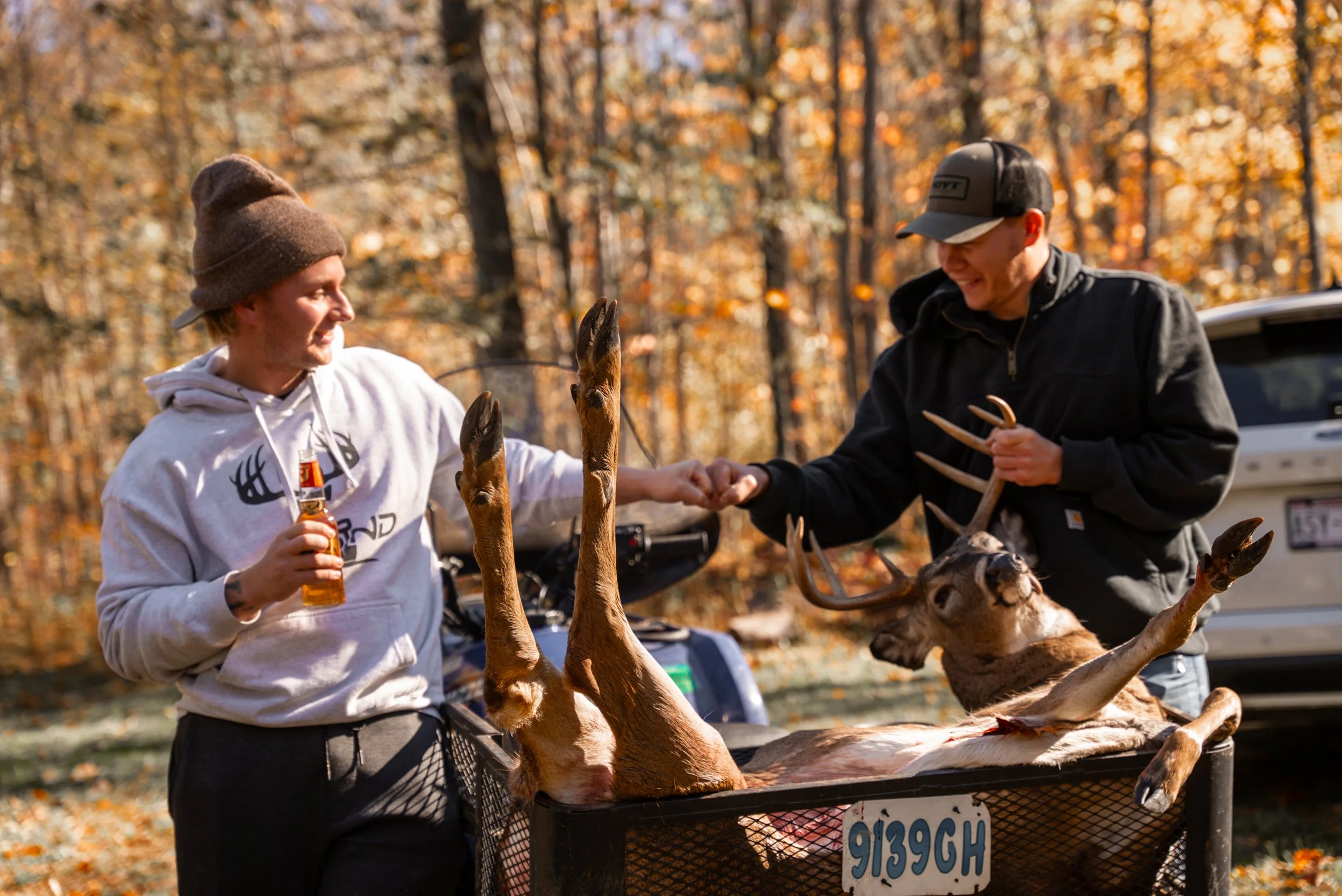 Two men in casual clothing and hats high-fiving over a harvested deer in a wooded area with fall foliage.