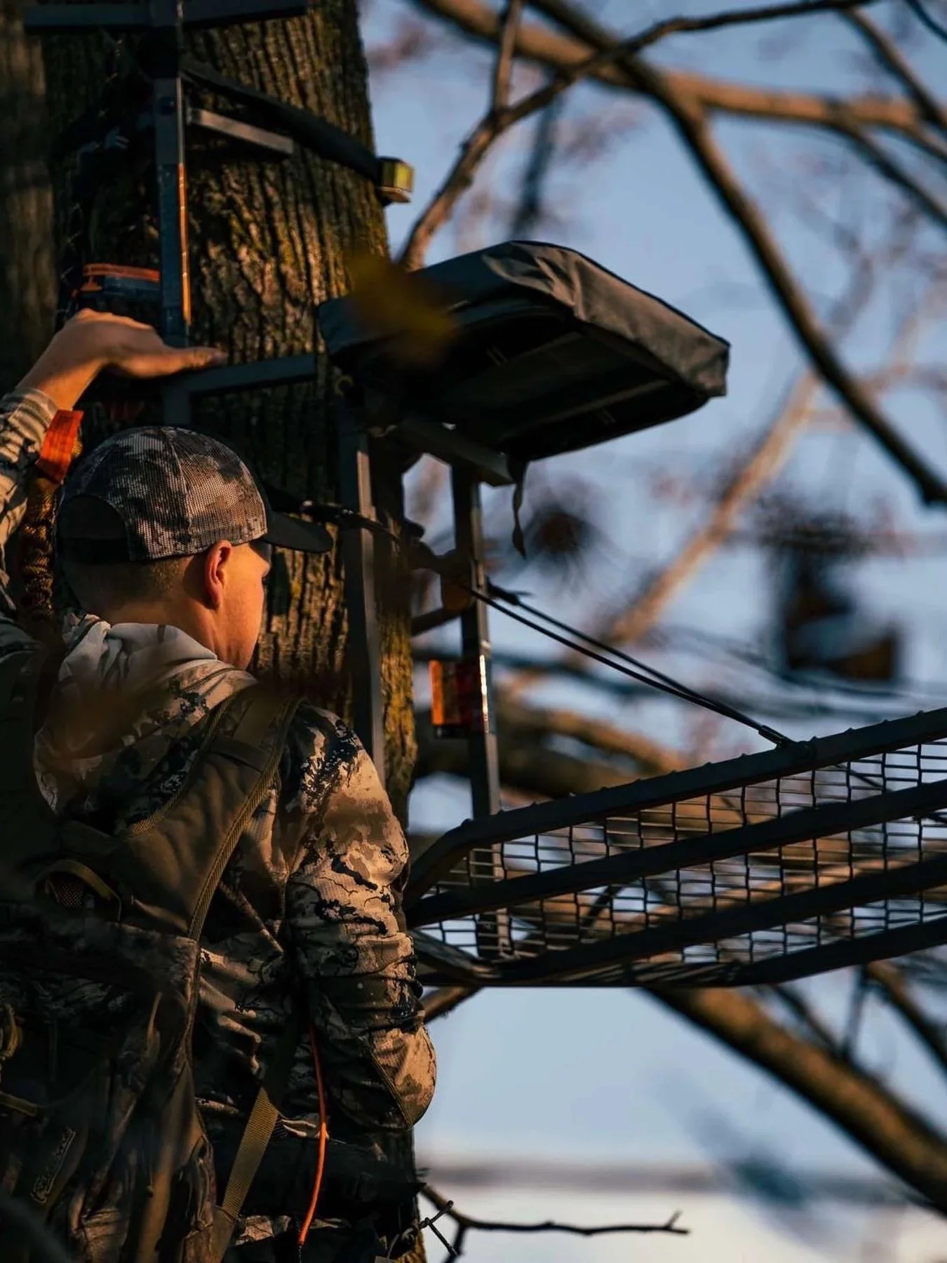 A man wearing a camouflage jacket and a camouflage baseball cap is climbing a metal hunting stand in a tree, with the sky and bare tree branches in the background during dusk or dawn.