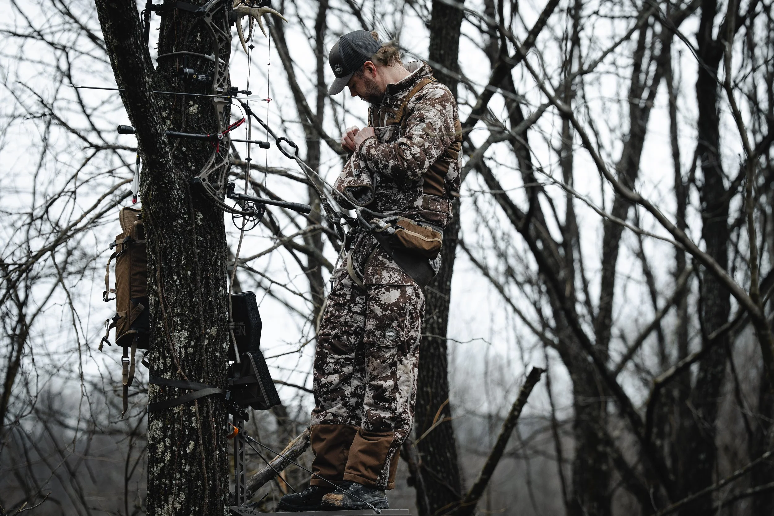 A man dressed in camouflage clothing is climbing a tree with a treestand and a compound bow attached to it in a forest during overcast weather.